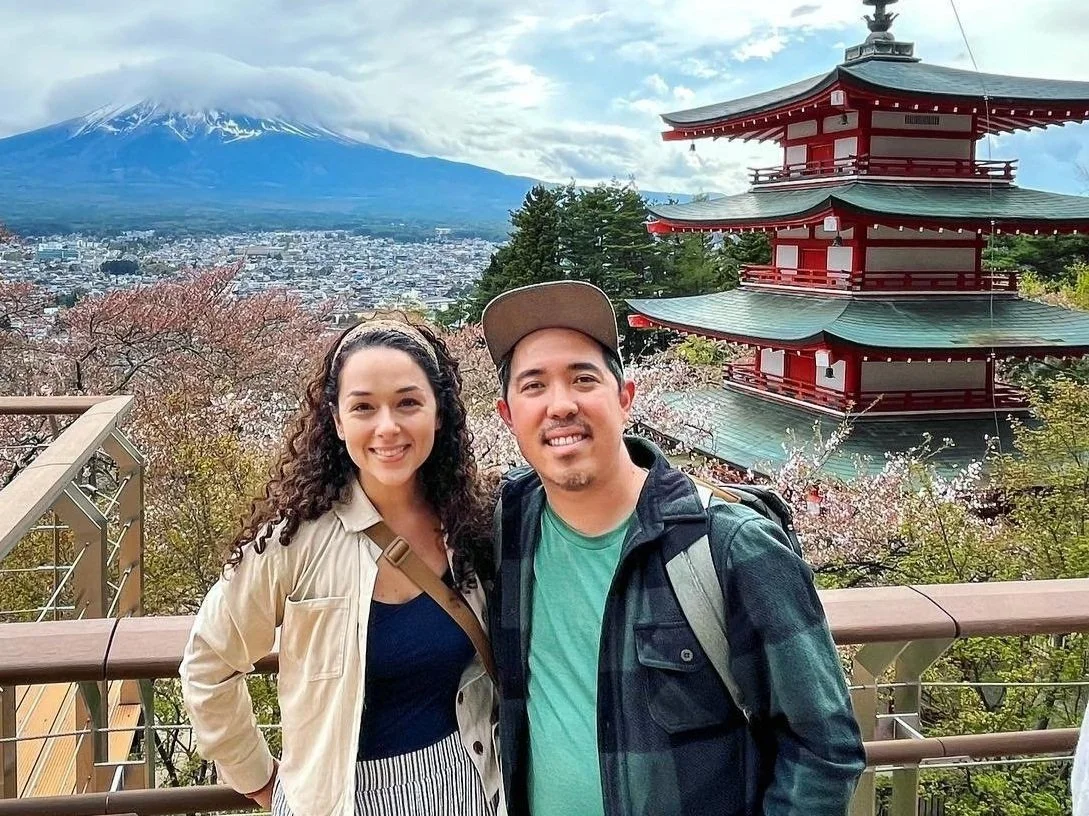 A smiling couple standing on a viewing platform with a scenic background of Mount Fuji, cherry blossoms, and a traditional Japanese pagoda