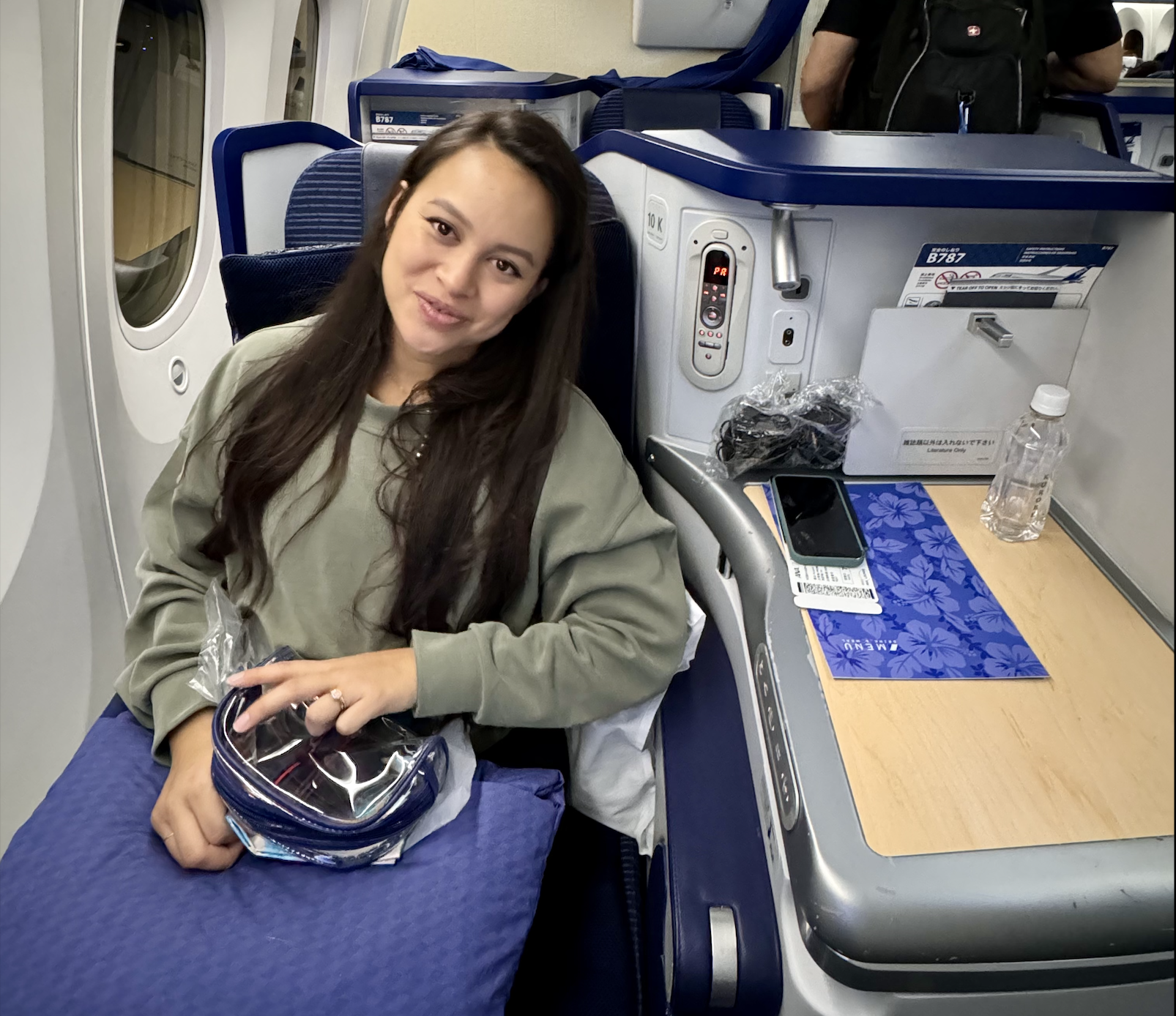 A woman sitting in an airplane seat by the window, smiling and looking at the camera. She has long dark hair and is wearing a khaki-colored jacket. On the tray table in front of her, there is a phone, a blue floral-patterned folder or magazine, a wat