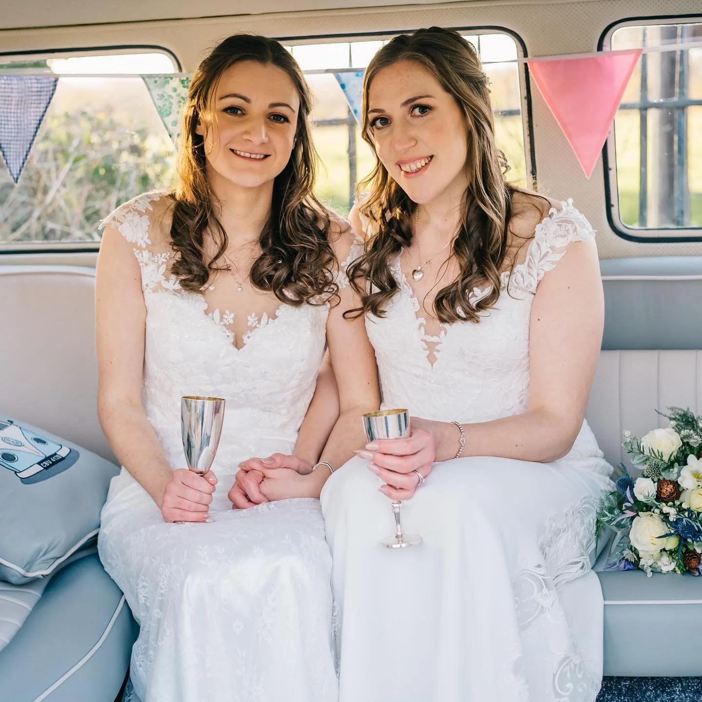 Two women wearing white wedding dresses sitting inside a decorated vehicle, holding champagne glasses, with colorful bunting in the background.