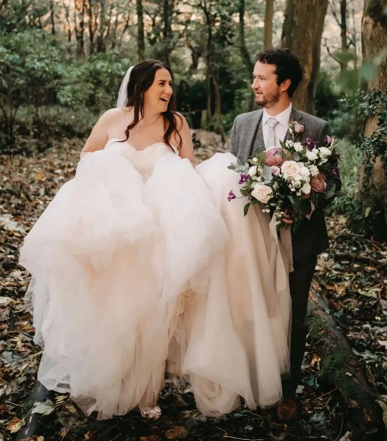 A bride and groom walking outdoors on a wooded trail, the bride in a white wedding dress and veil, the groom in a gray suit holding a bouquet of pink, white, and purple flowers, smiling at each other.