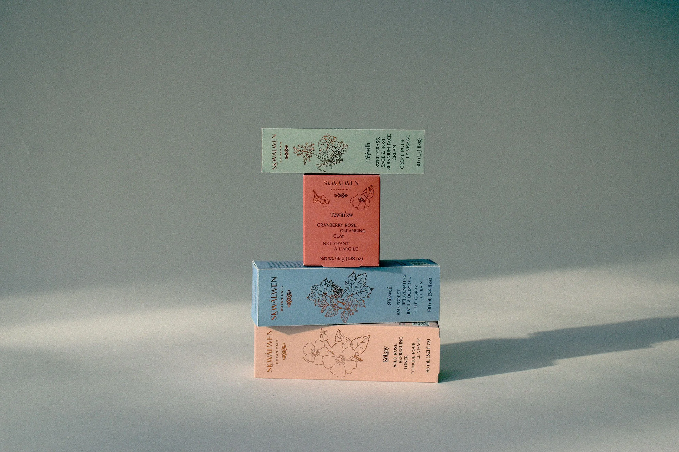 Stack of Skwalwen skincare product boxes in pastel colors, arranged on a shadowy white background. Each box has line art of a botanical drawing drawn by Sydney Mak.