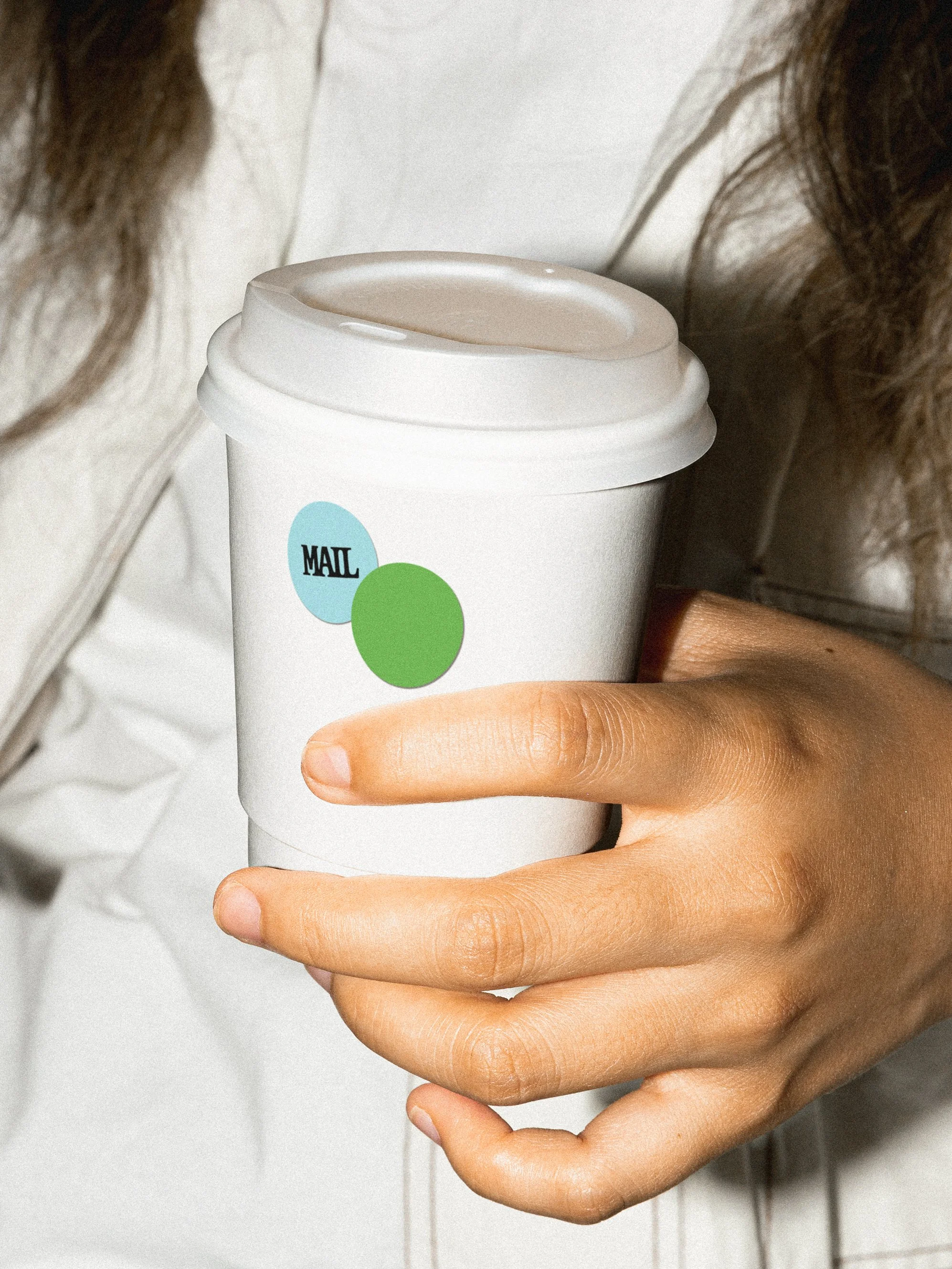 A person holding a white disposable coffee cup with a lid, featuring two circle stickers, one blue with 'MAIL' on it in black, and one green. Modular stickers.