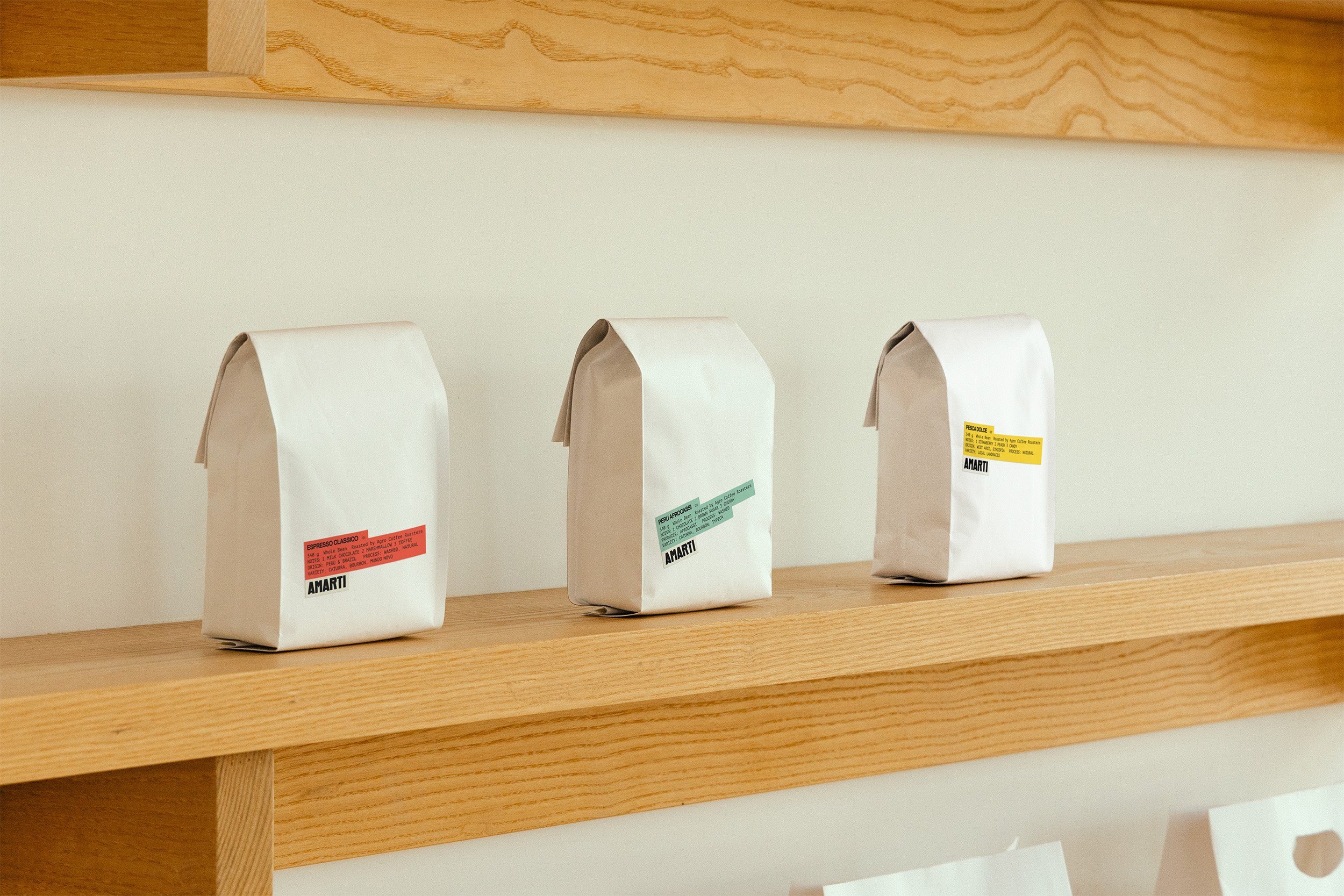 Three white coffee bags with colored labels on a wooden shelf against a white wall.