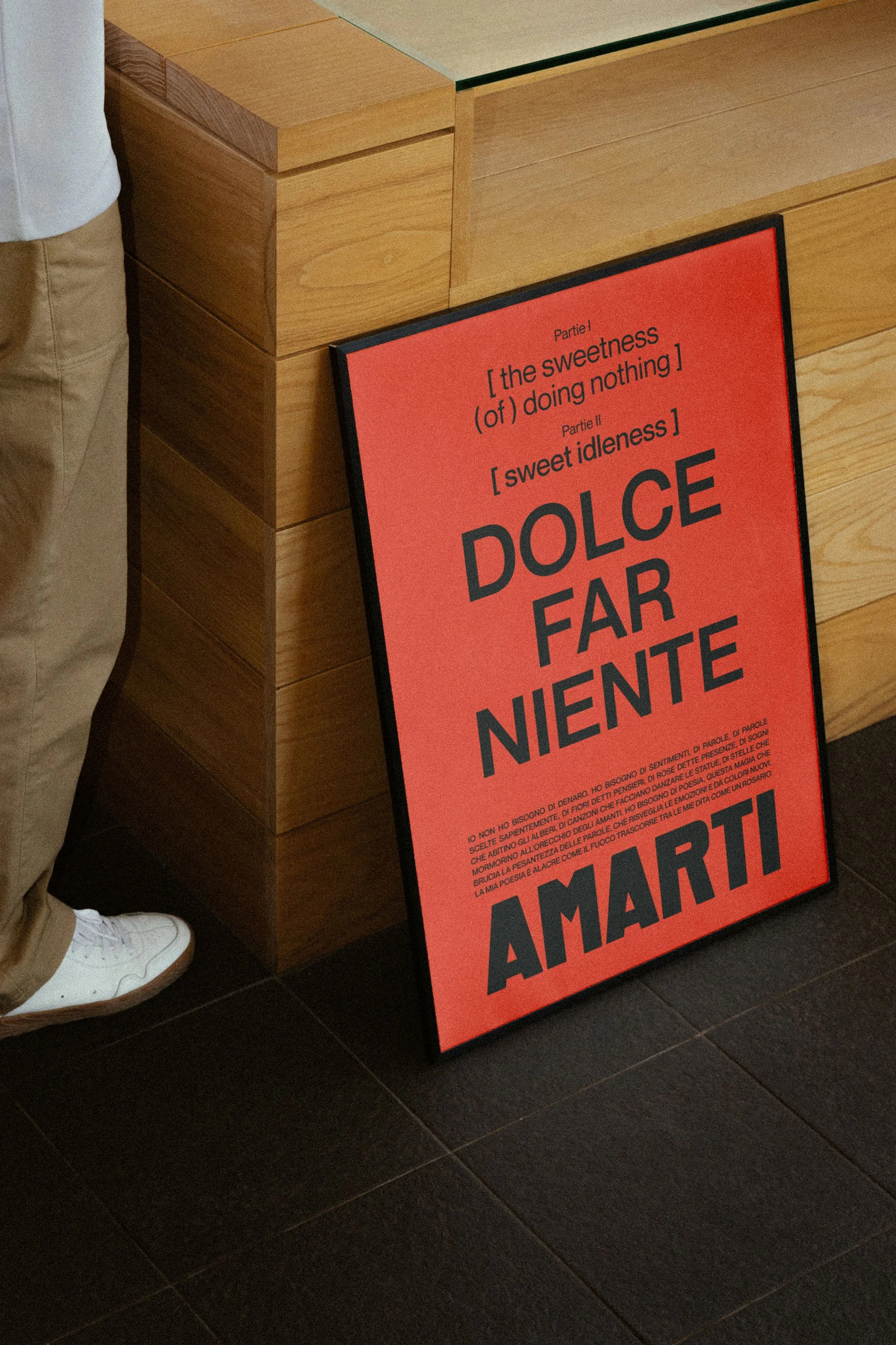 A red poster with black bold text stands on the dark tiled floor, next to a wooden counter and a person wearing khaki pants and white sneakers partially visible.