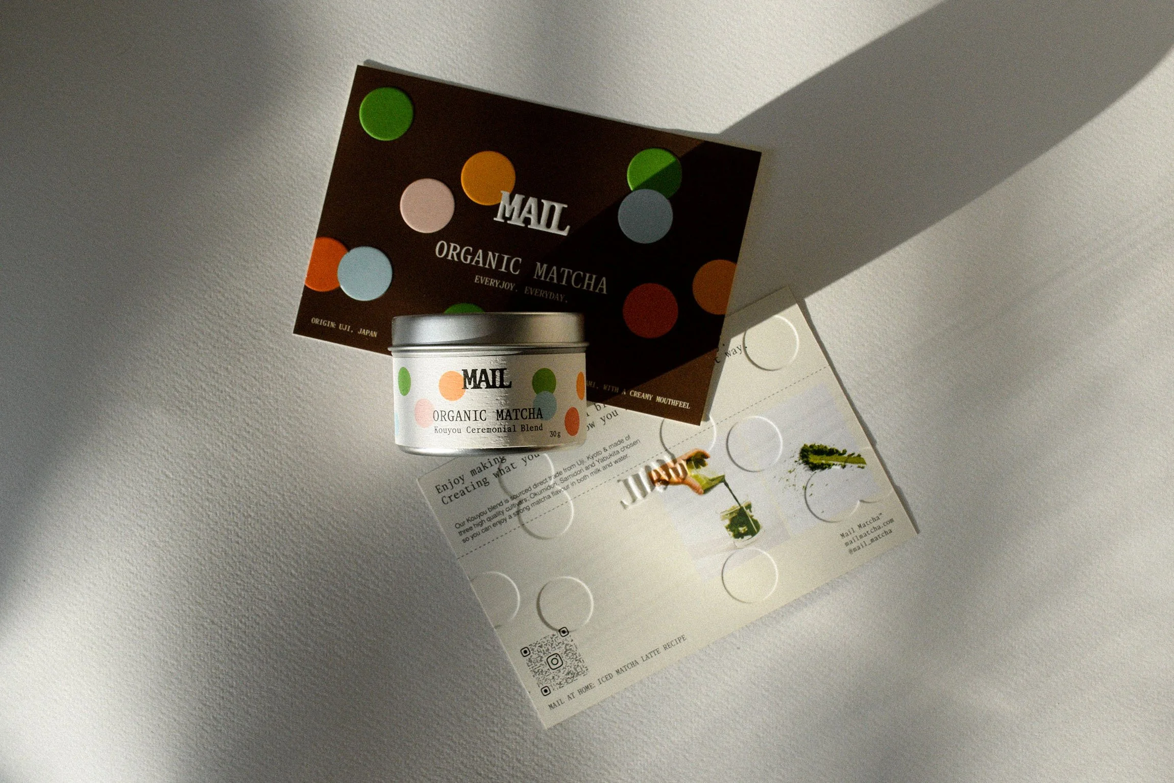 A small white container labeled 'Mail Organic Matcha' sitting on a sheet of paper with informational text, with a brown informational card behind it. The paper has circular cutouts and a small illustration of a tea leaf. The scene is lit with a shadow cast on the right side.