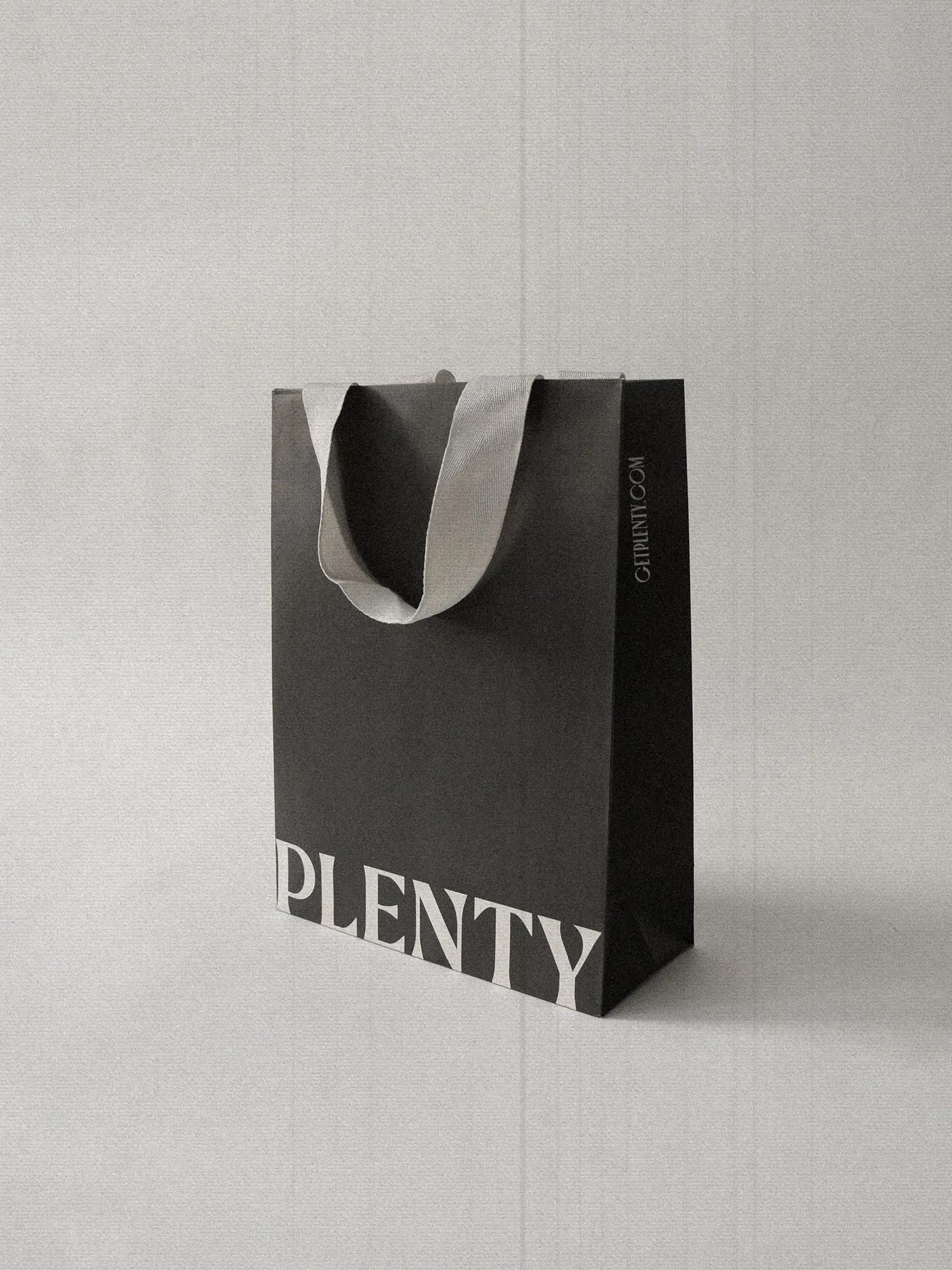 A black and white image of a structured black gift bag with white handles, displaying the word 'PLENTY' in large white letters on the front, set against a light background.