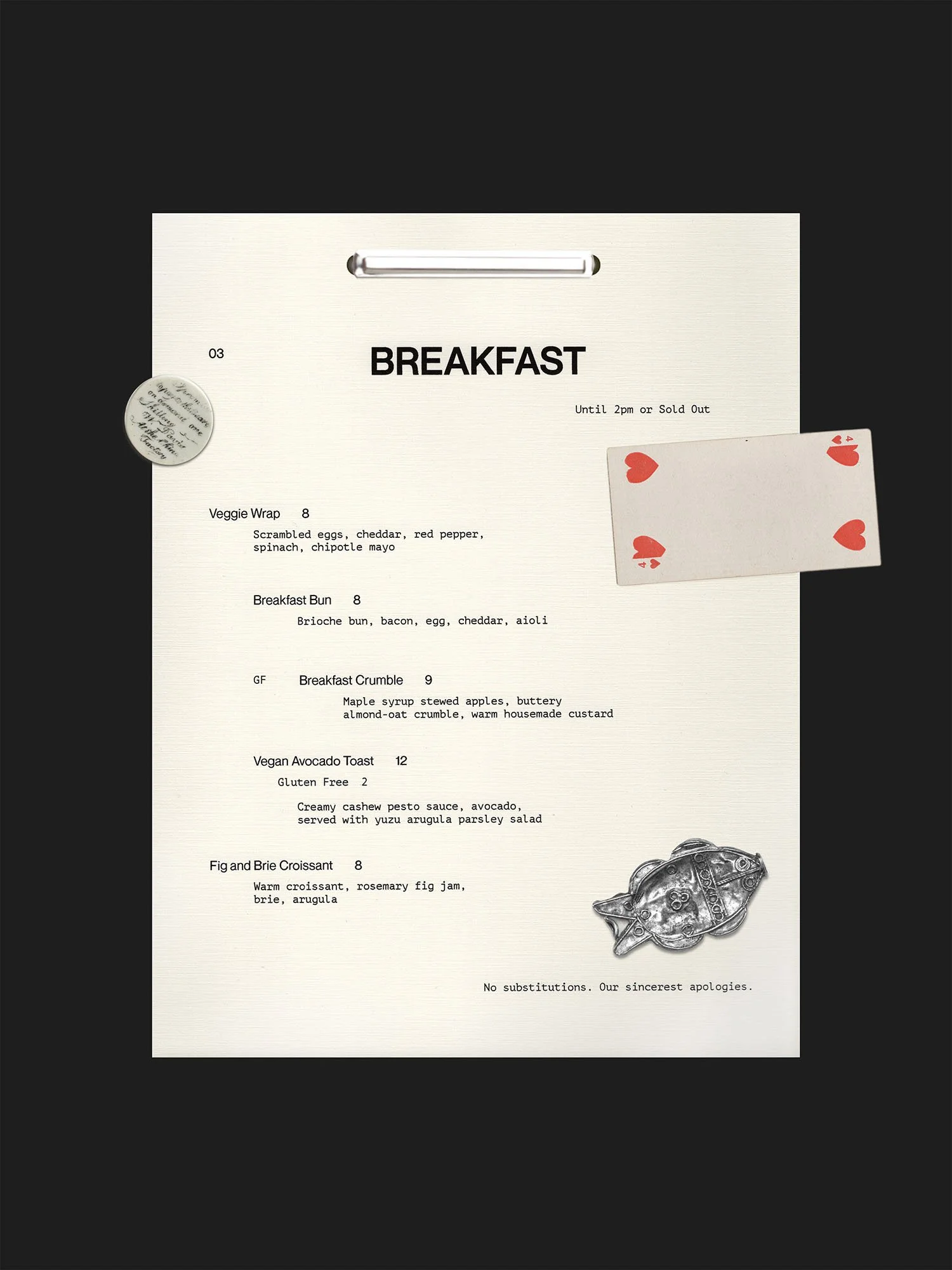 A breakfast menu on white paper with black text, featuring items like veggie wrap, breakfast bun, breakfast crumble, vegan avocado toast, and fig and brie croissant, placed on a black background with a decorative fish design and a card with red hearts attached.