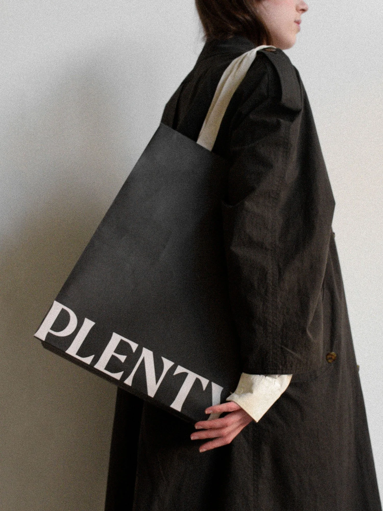 A person wearing a black coat carrying a large structured shopping bag with the word 'PLENTY' printed large in white. The person is standing against a neutral background.