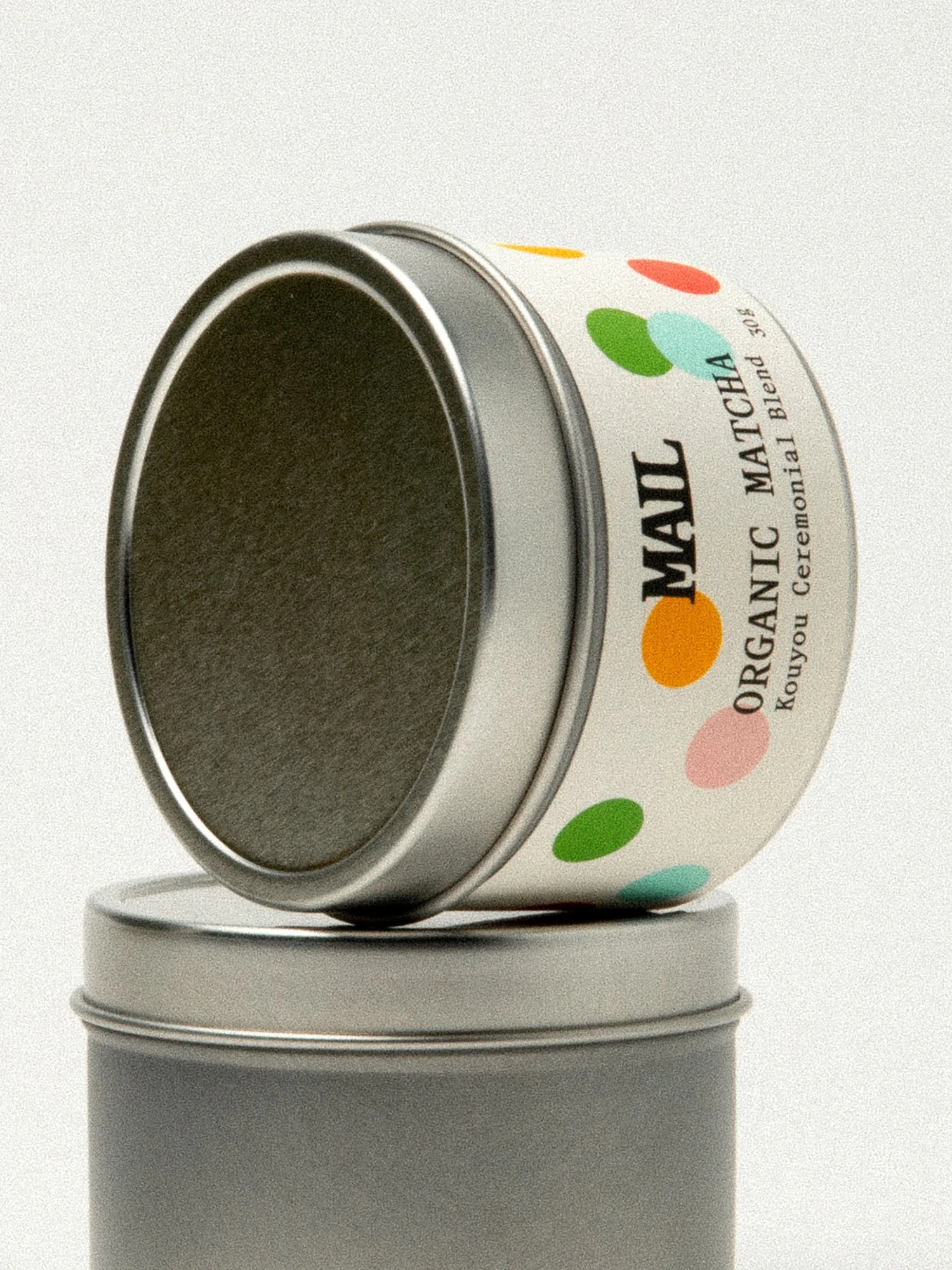 Close-up of a round metal tin with colorful polka dots, labeled as organic matcha, arranged on a plain surface with a white background.