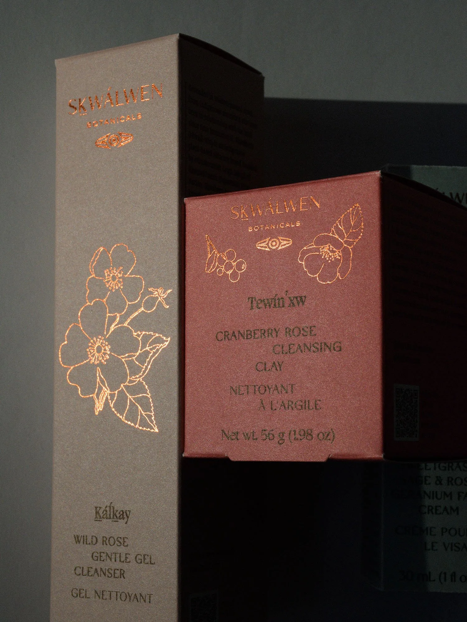 Boxes of Skwalen Botanicals skincare products, including a wild rose gel cleanser and a cranberry rose cleansing clay, with floral and berry designs on the packaging.