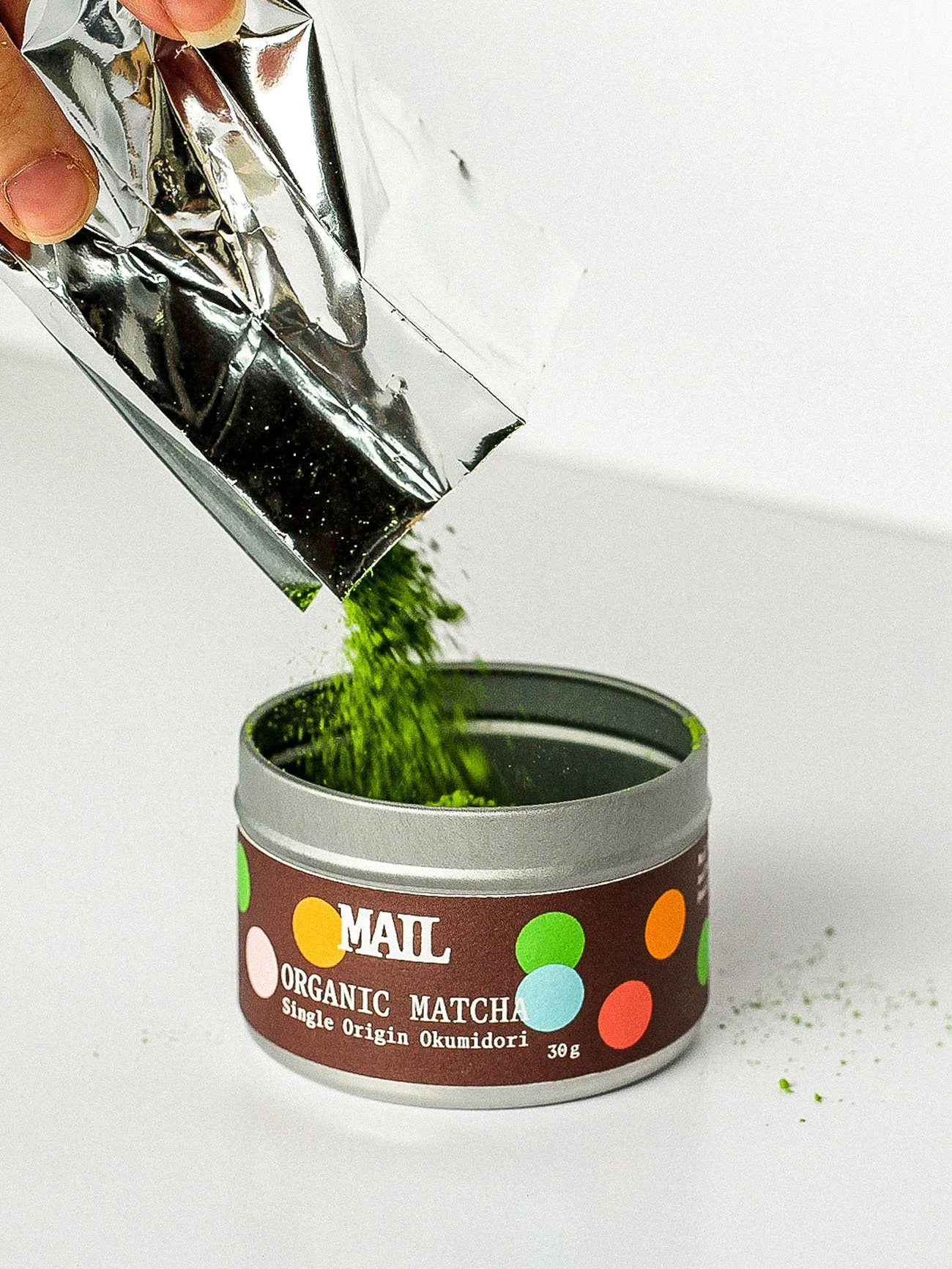 Green matcha powder being poured from a silver packet into a round container labeled "Organic Matcha" on a white surface.