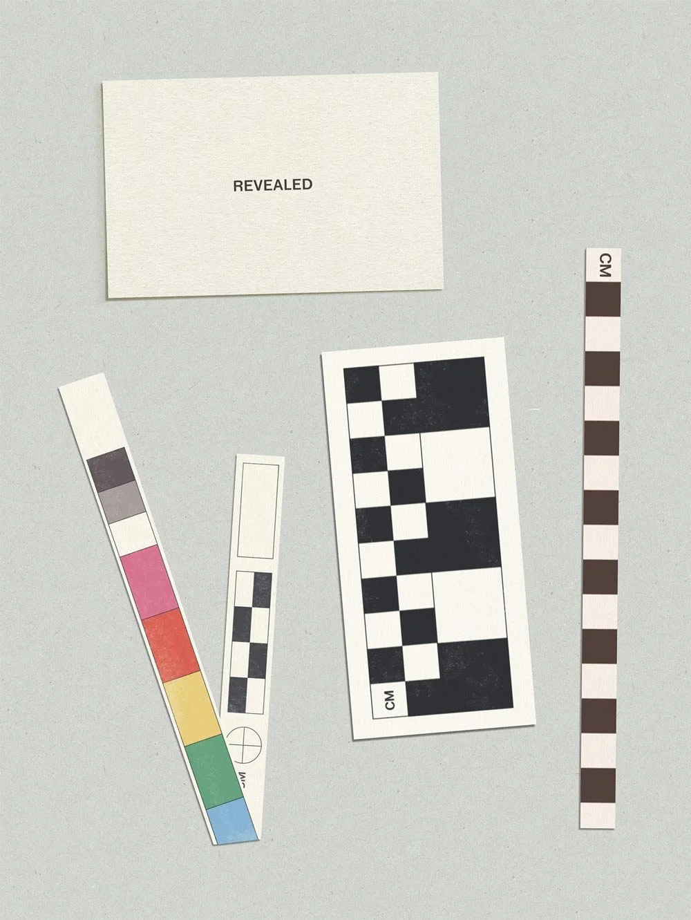 Photo of three bookmarks inspired by artifact cataloging tools like color and black-and-white calibration charts, and a card with the word 'REVEALED' on it, arranged on a gray surface.
