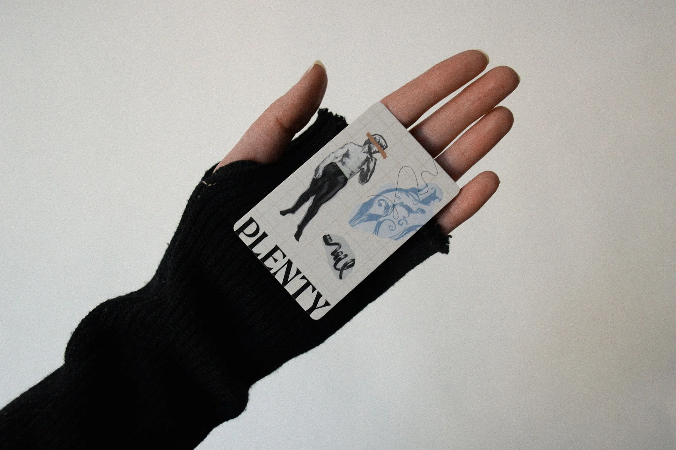A hand wearing a black fingerless glove holding a small card with a black and white drawing of an old man on the front, featuring the word 'PLENTY' in bold letters, against a plain light-colored background.