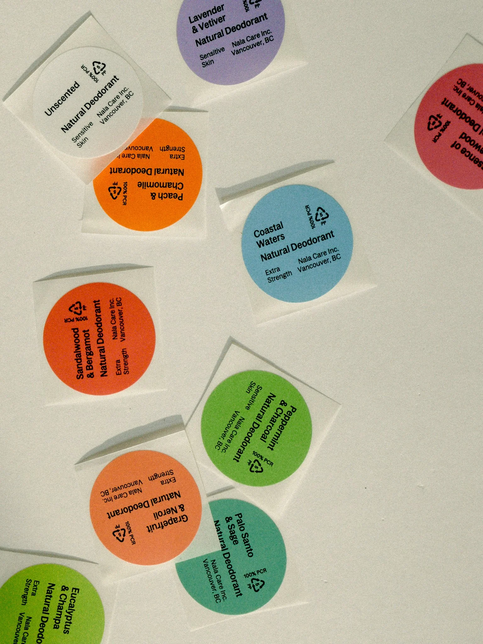 Multiple circular labels on a white surface, each with product information for natural deodorants in different colors including purple, white, orange, blue, red, green, and teal.
