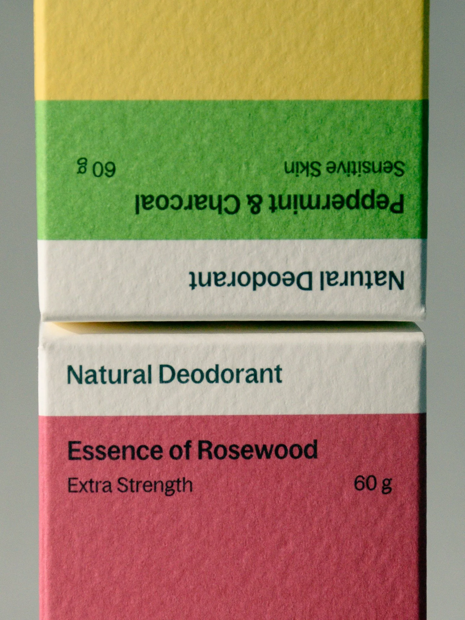 Close-up of two boxes of natural deodorant; one with white and pink packaging labeled 'Essence of Rosewood Extra Strength 60 g' and the other with green and yellow packaging labeled 'Peppermint & Charcoal Sensitive Skin 60 g'.