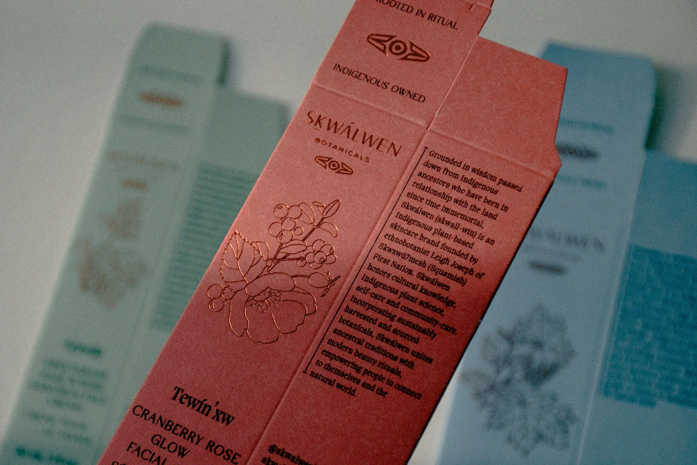 Close-up of a pink skincare product box with gold foil details, featuring text about indigenous ownership and plant-based ingredients, with other skincare boxes in pastel colors blurred in the background.