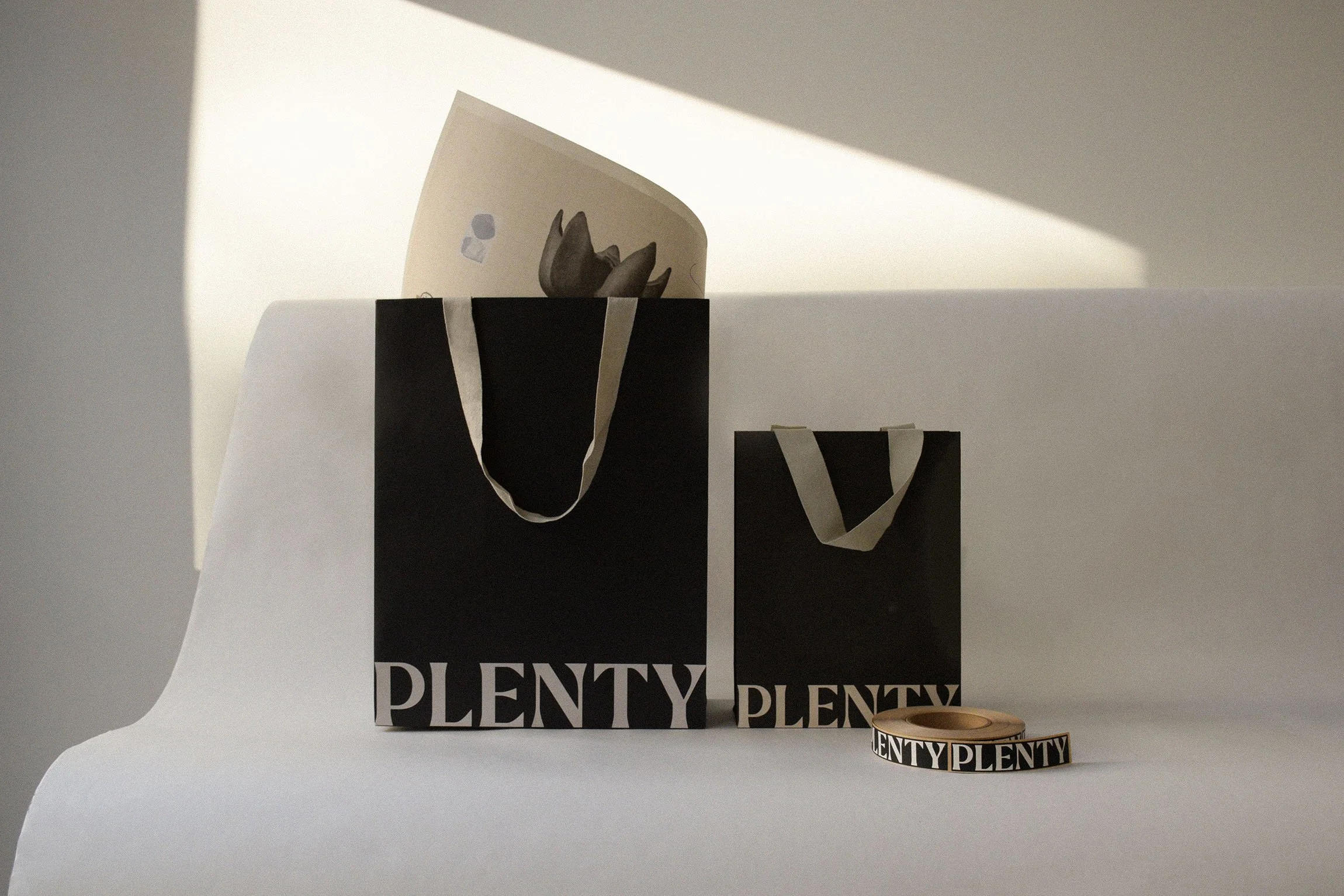 Black shopping bags with white handles and the word 'PLENTY' printed on them, one larger and one smaller, along with a roll of black rectangle textured stickers reading 'PLENTY' in white. All on a white surface against a white background.