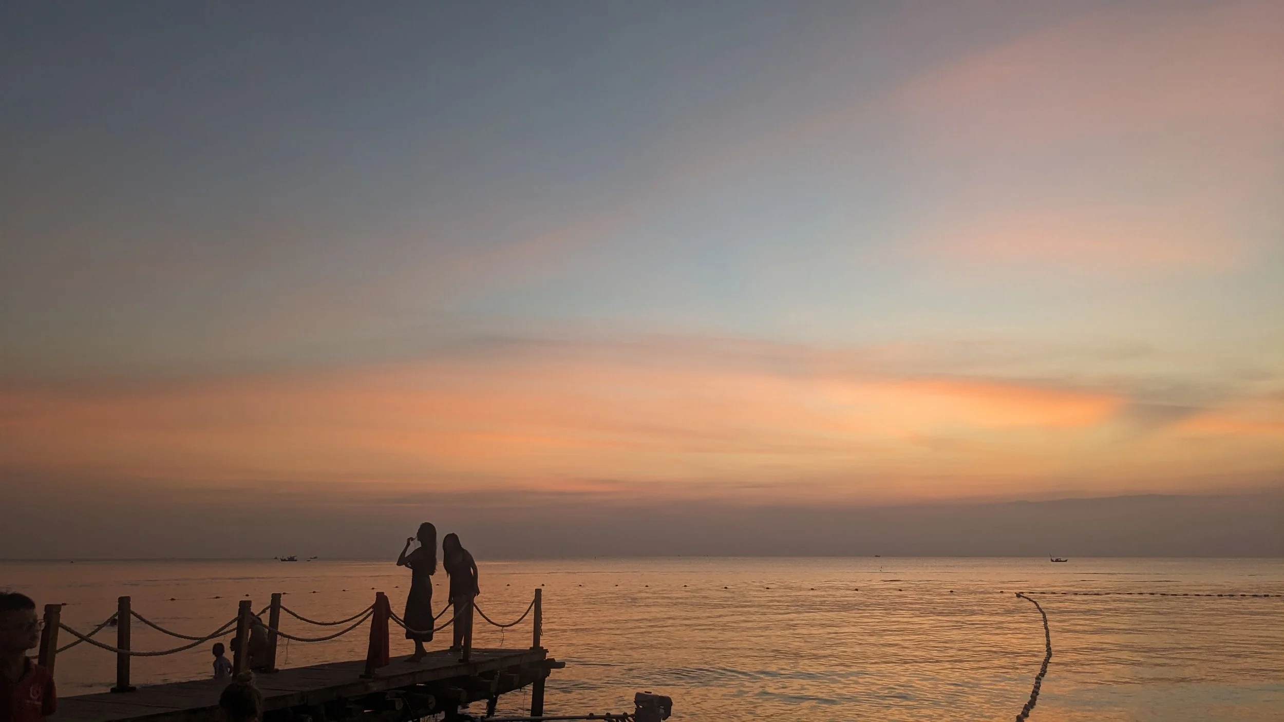 Silhouettes of people on a dock watching the sunset over the ocean with colorful sky and distant boats.