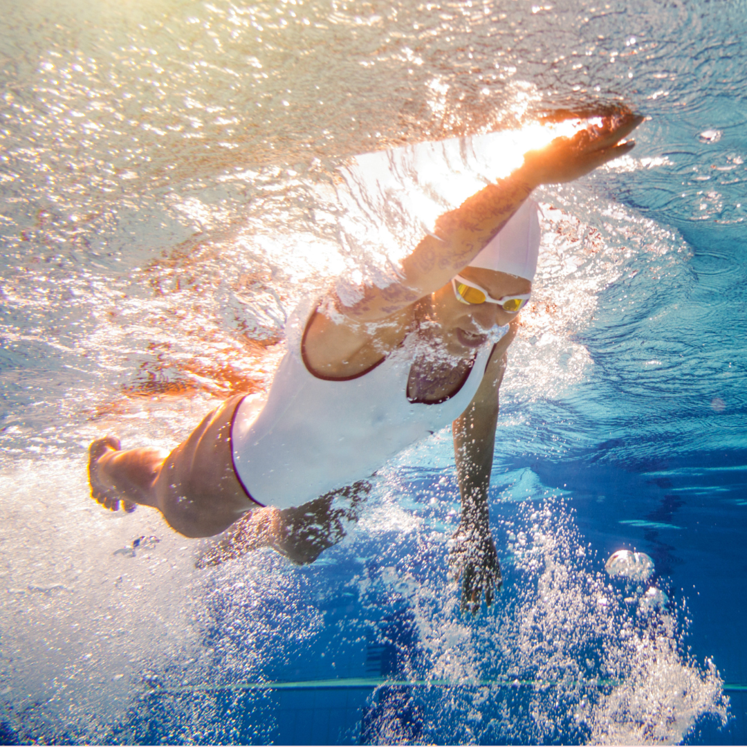 Swimmer in a pool wearing a white swim cap and yellow goggles, diving underwater with a splash and bubbles around.