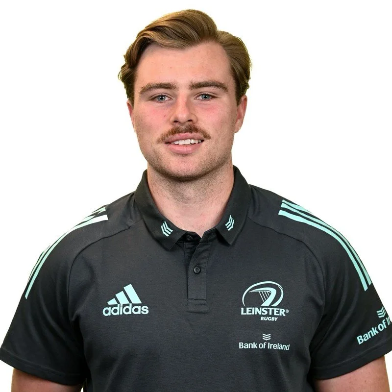 A young man with light brown hair, a mustache, and a slight smile, wearing a black rugby jersey with blue accents and various logos, including Adidas, Leinster Rugby, and Bank of Ireland.