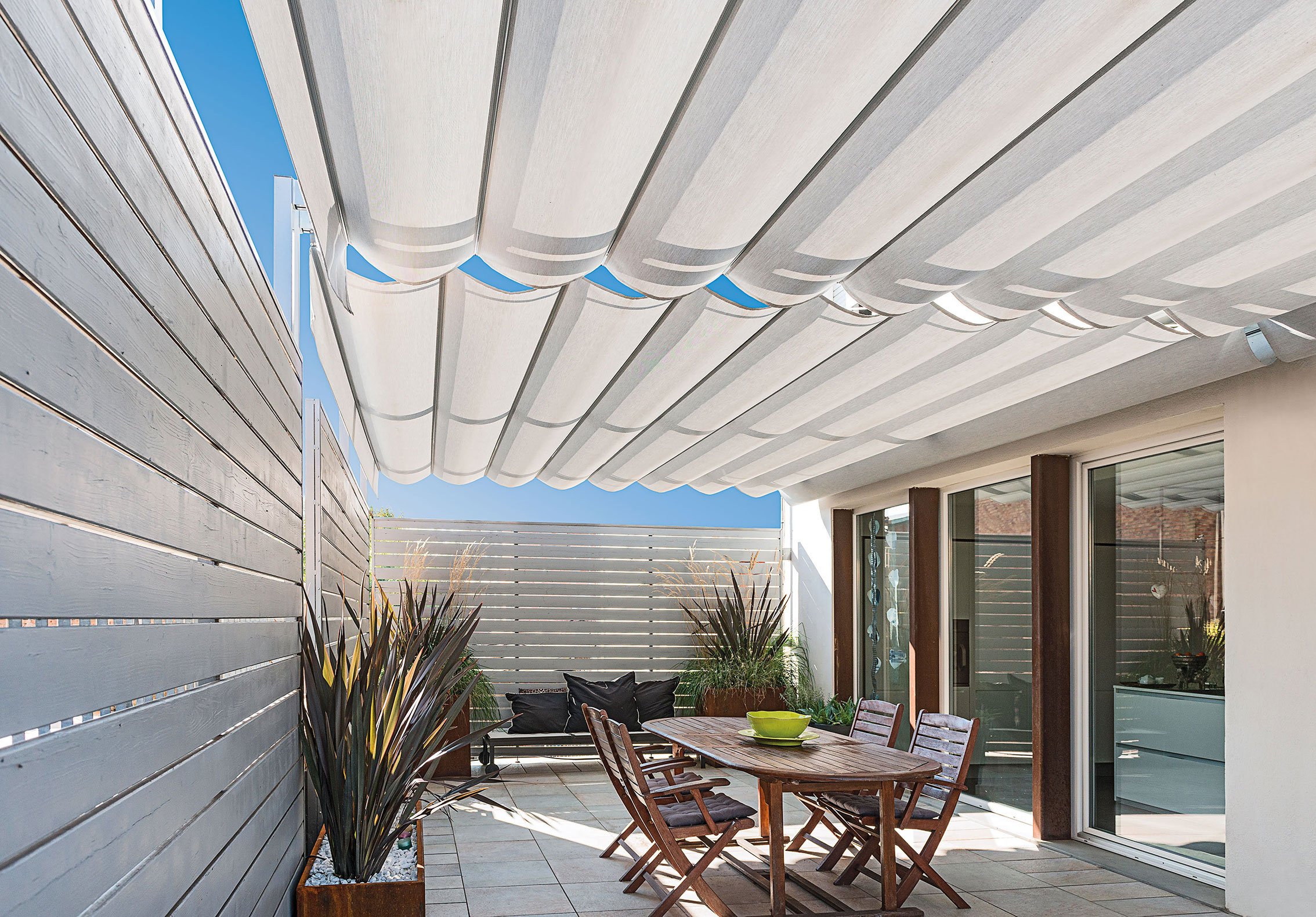 Terrazza con tavolo e sedie in legno, piante ornamentali, tende parasole e recinzione in legno.