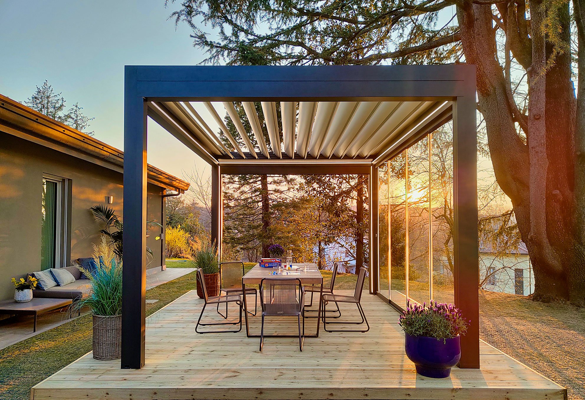Pergola su terrazza con tavolo e sedie, ambiente esterno con piante e tramonto.