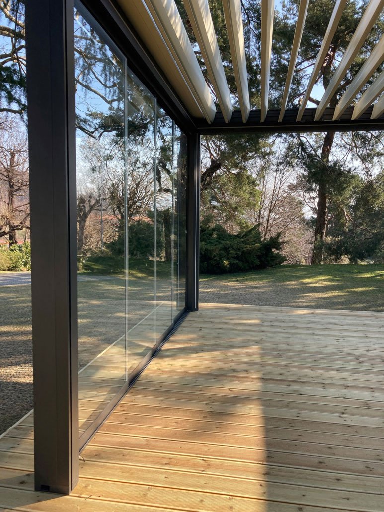 Terrazza con pavimento in legno, chiusa con pareti di vetro e coperta da lamelle frangisole, circondata da alberi.