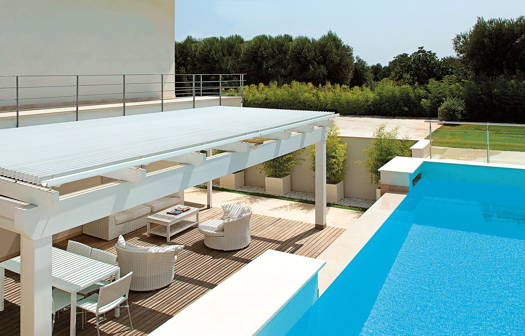 Patio con piscina blu, mobili da esterno bianchi e pergola con copertura. Alberi e vegetazione sullo sfondo.