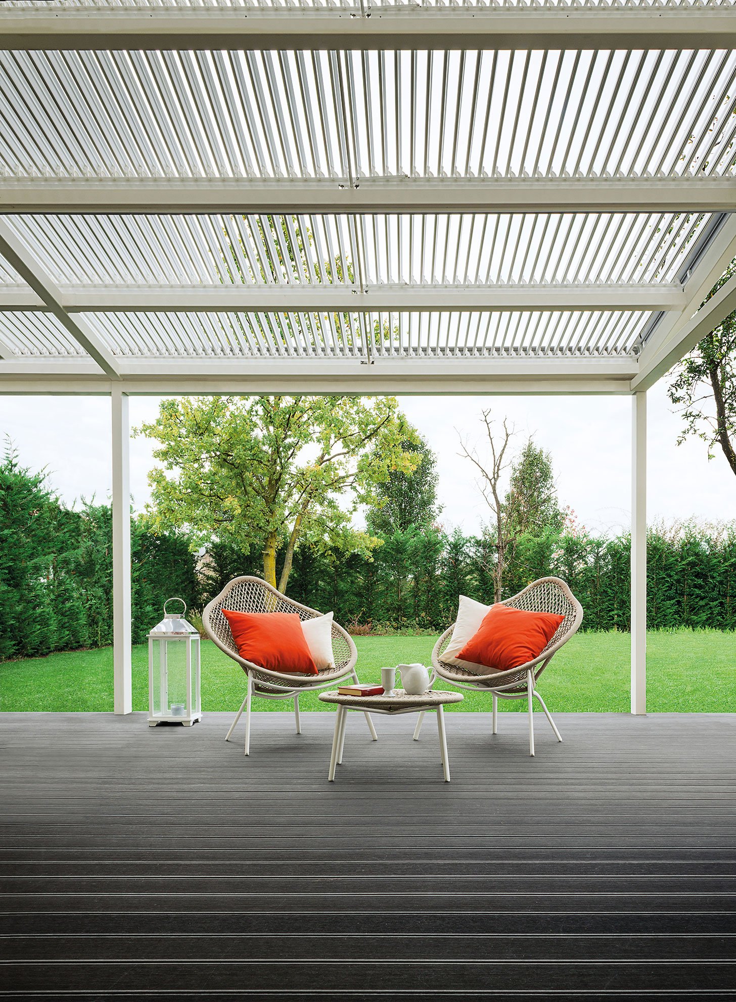 Terrazza con due sedie di vimini con cuscini arancioni, un tavolino con tazze e una lanterna, sotto una pergola, con giardino sullo sfondo.