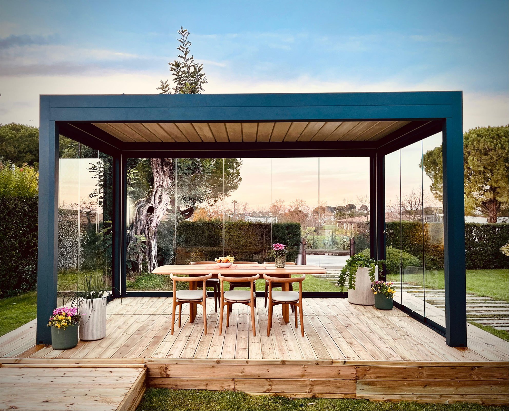 Gazebo moderno con struttura in metallo e pannelli di vetro, tavolo di legno con sedie, piante in vaso, sfondo di giardino e alberi, cielo sereno.