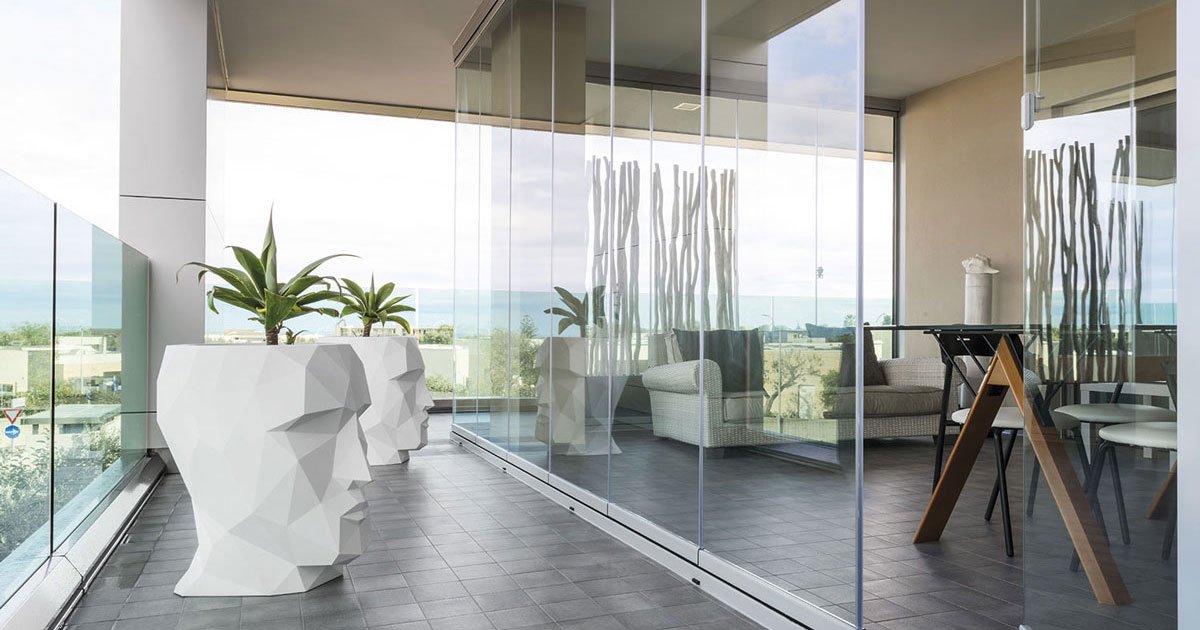 Vista di una terrazza moderna con pavimento in piastrelle grigie, vasi bianchi a forma di testa contenenti piante, un'area salotto con divano e tavolino, e ampie vetrate che danno su un panorama esterno.
