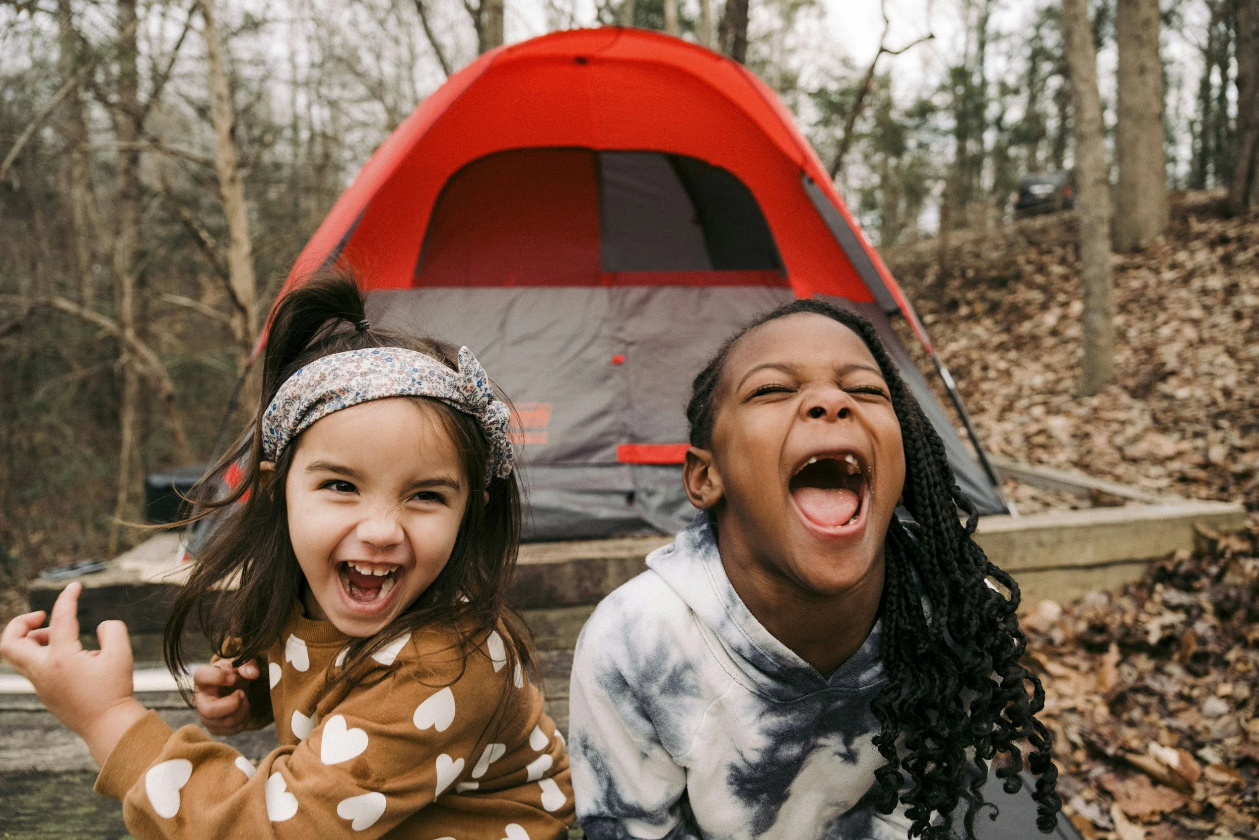 Two young girls smiling and laughing in front of a red camping tent in a wooded area.