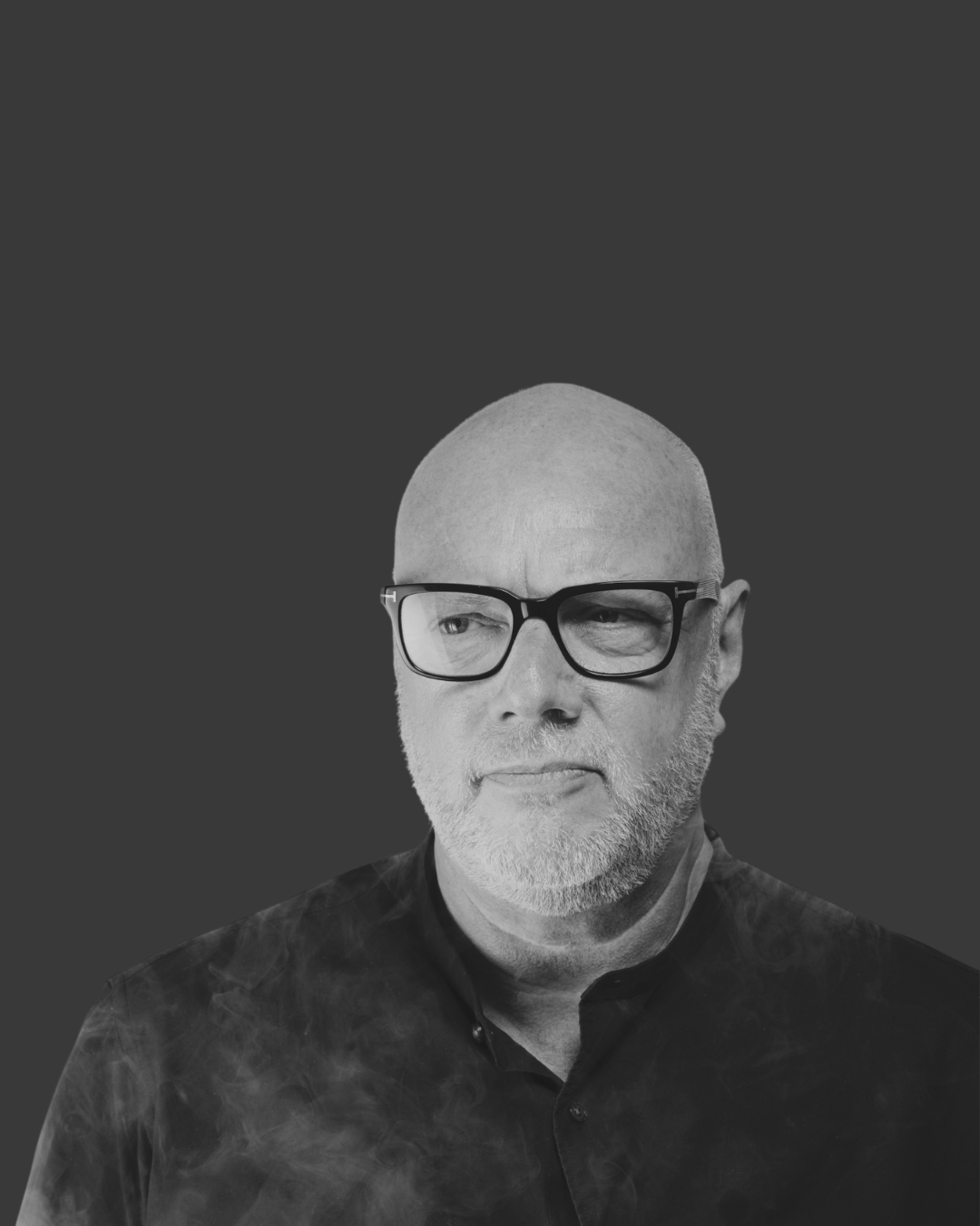 Black and white portrait of an older man with a serious expression, bald head, beard, wearing glasses, and a dark collared shirt, against a plain dark background.