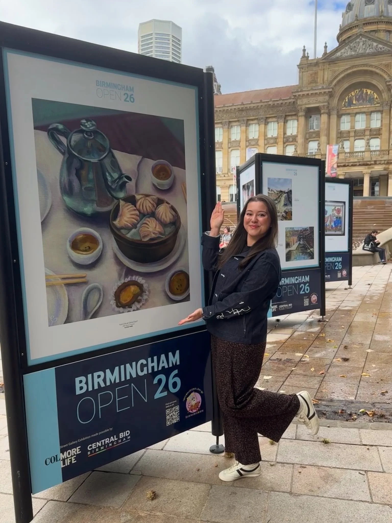 a photo of the artist emily peddle standing in front of a painting of dim sum, displayed in the Birmingham Open art exhibition