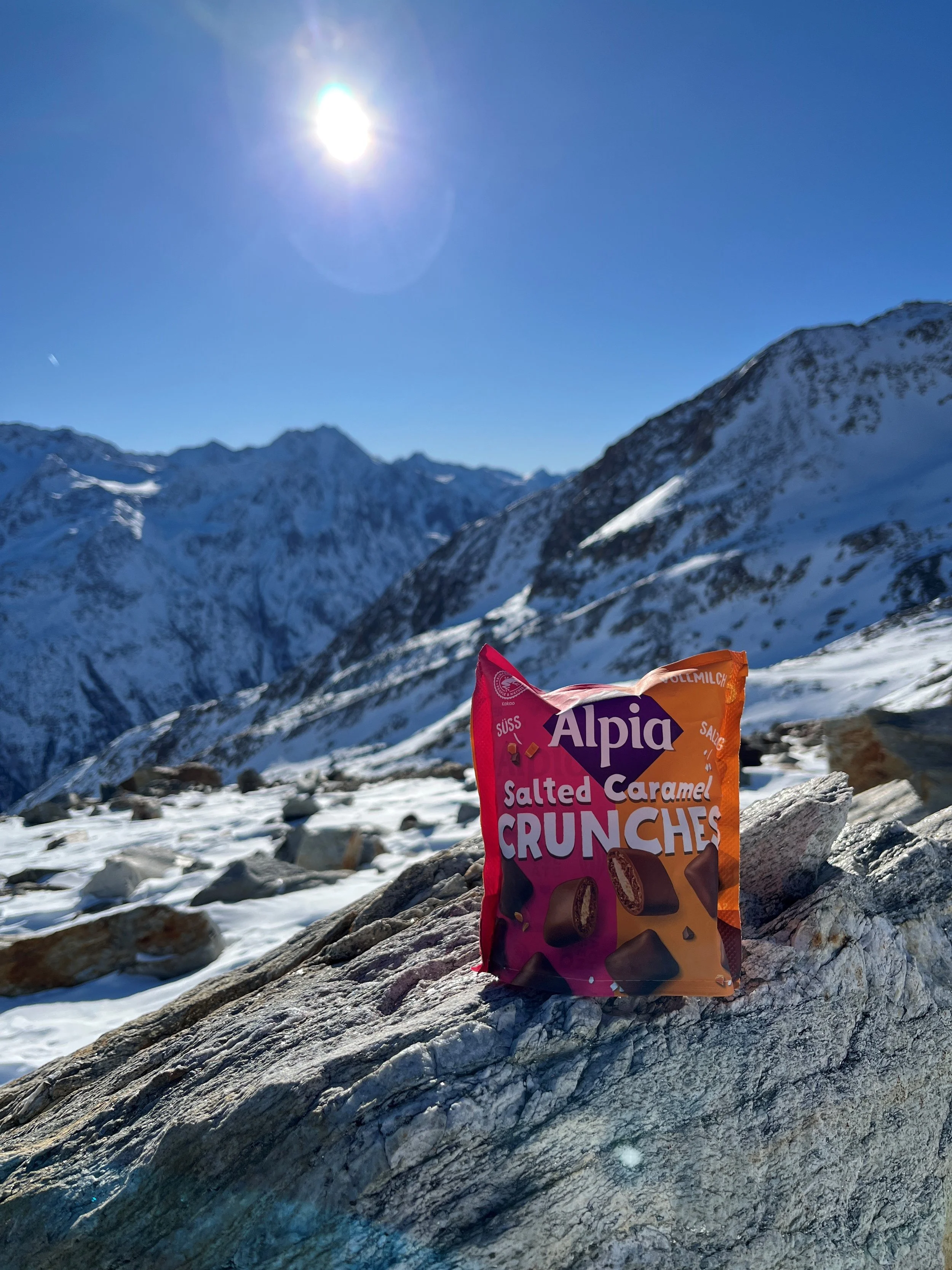 Alpia Salted Caramel Crunches – salzig-süßer Schokoladensnack in winterlicher Schneelandschaft mit Sonnenschein auf dem Berg.