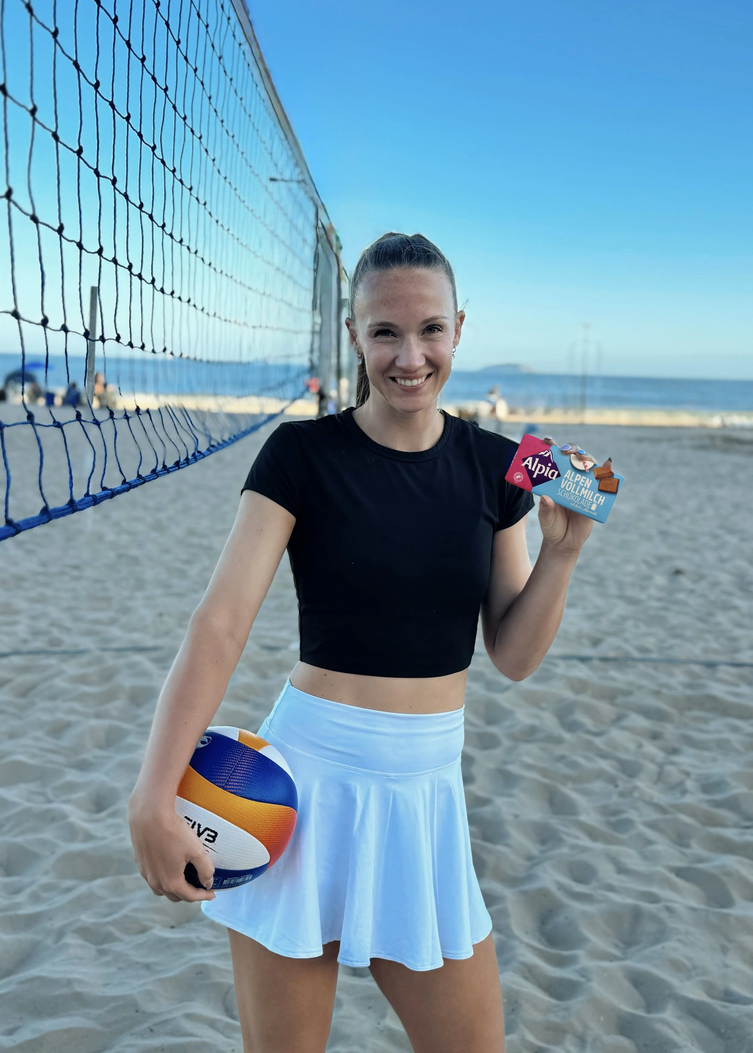 Beachvolleyball-Spielerin Sandra steht mit einem Volleyball am Strand neben dem Netz und präsentiert zwei Alpia Vollmilch Schokoladentafel.