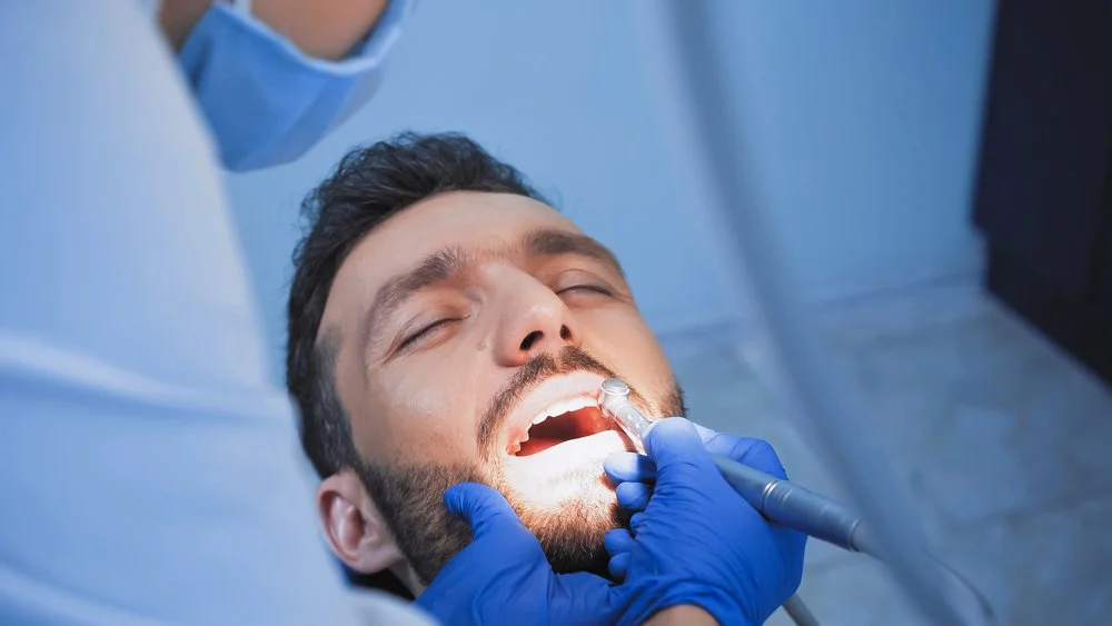 A man lying back with his mouth open while a dentist checks his teeth using a dental mirror, the dentist wearing gloves and a face mask.
