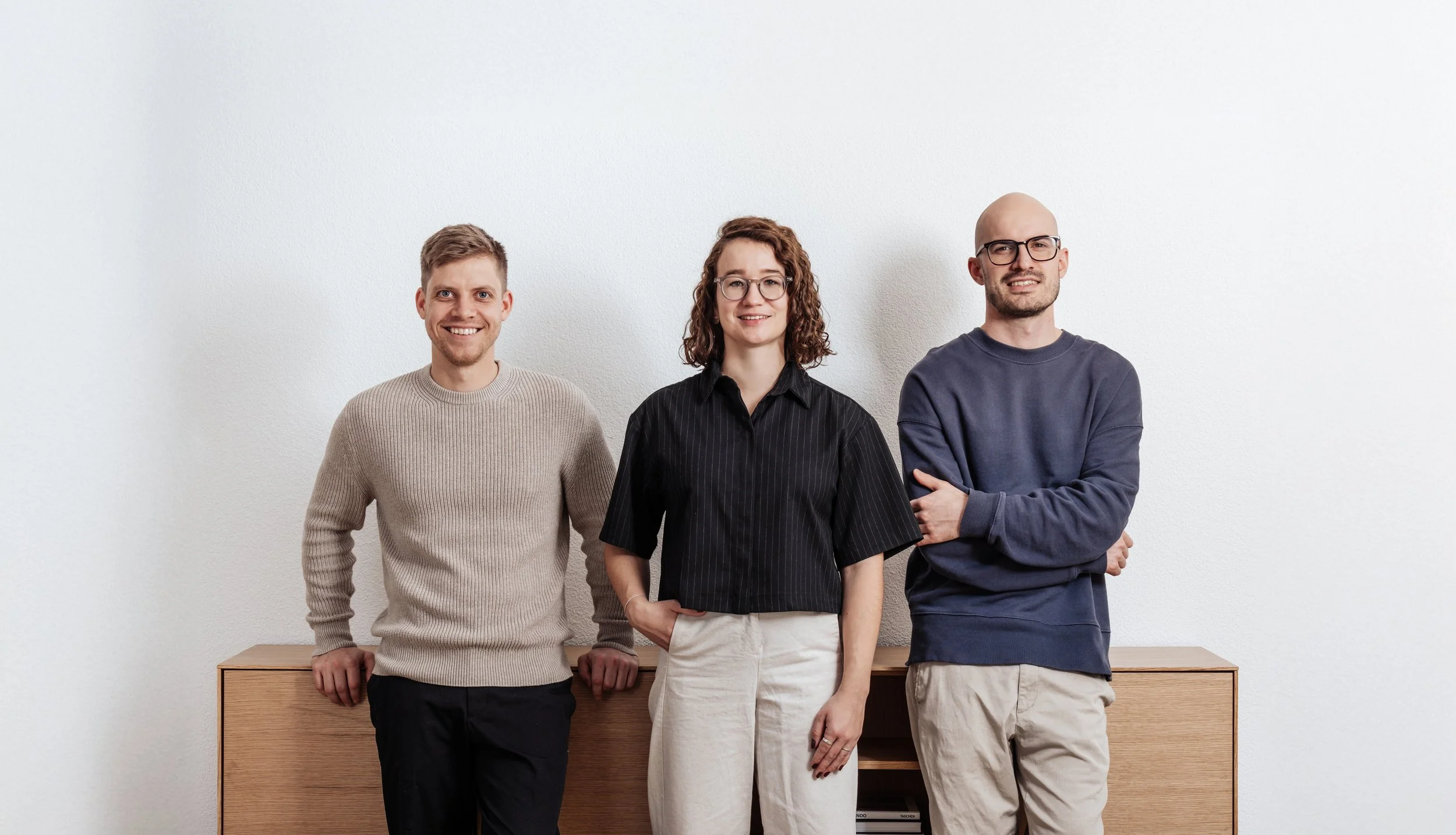Drei Personen (Benjamin Schalk, Noëmi Burkhalter und Florian Kech) stehen vor einer weißen Wand, lächeln und haben die Hände in den Taschen oder verschränkt. Im Hintergrund ist ein Holzschrank sichtbar.