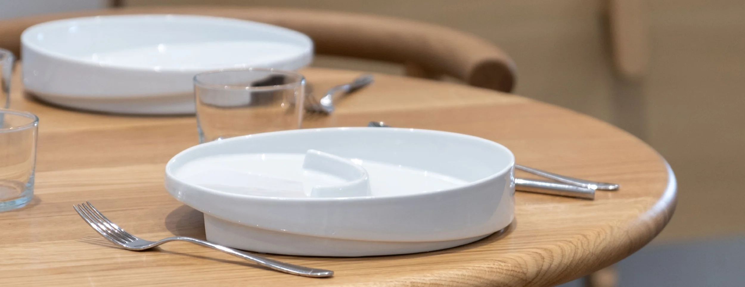 Leeres weißes Teller mit erhöhtem Tellerrand und Unterteilung auf einem Holztisch, daneben ein Messer und ein Gabel, im Hintergrund weitere universelle Teller mit Randerhöhung, Schräge und rutschfestem Boden.