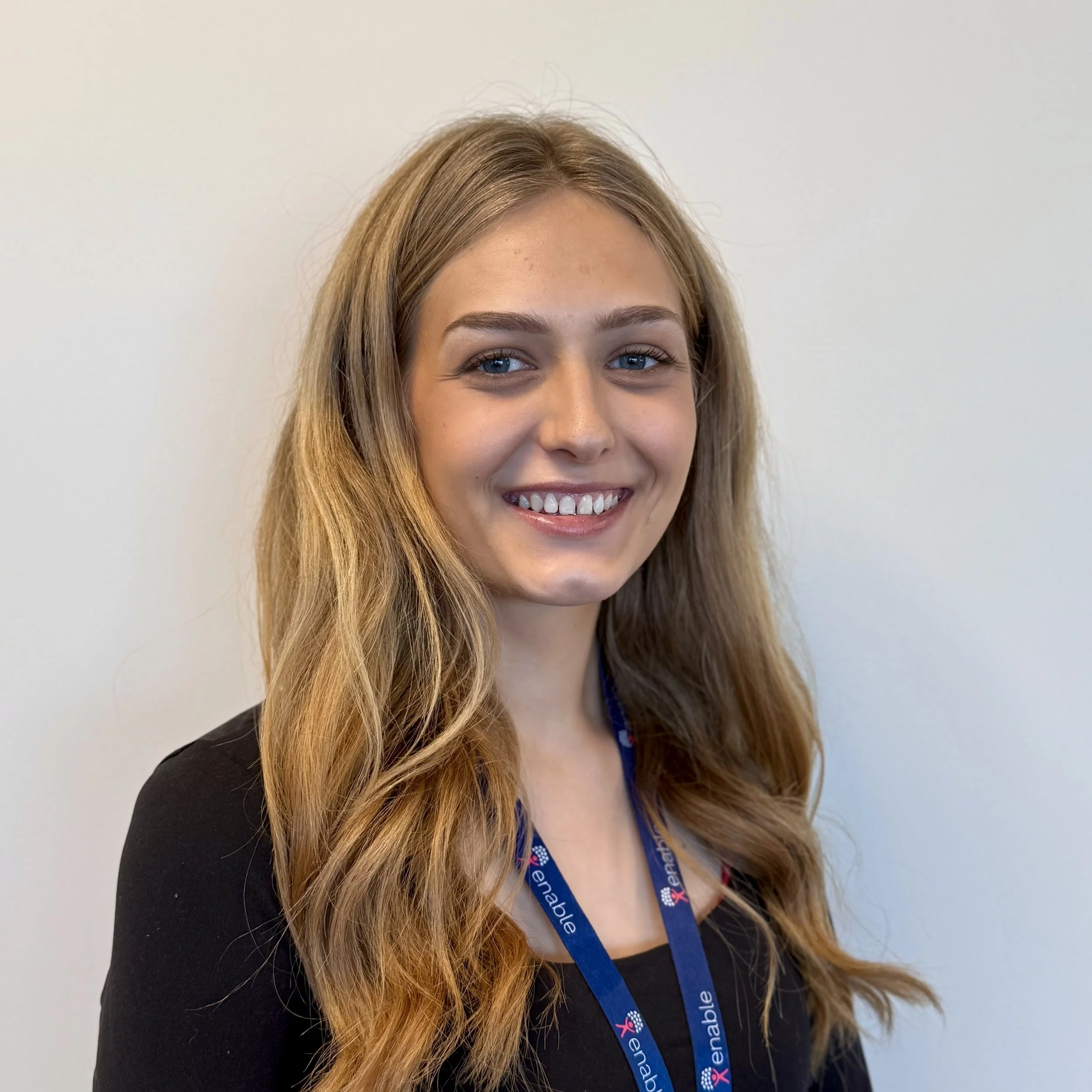 A woman with long dark blonde hair smiling against a plain background with a black top and blue 'Enable' lanyard.