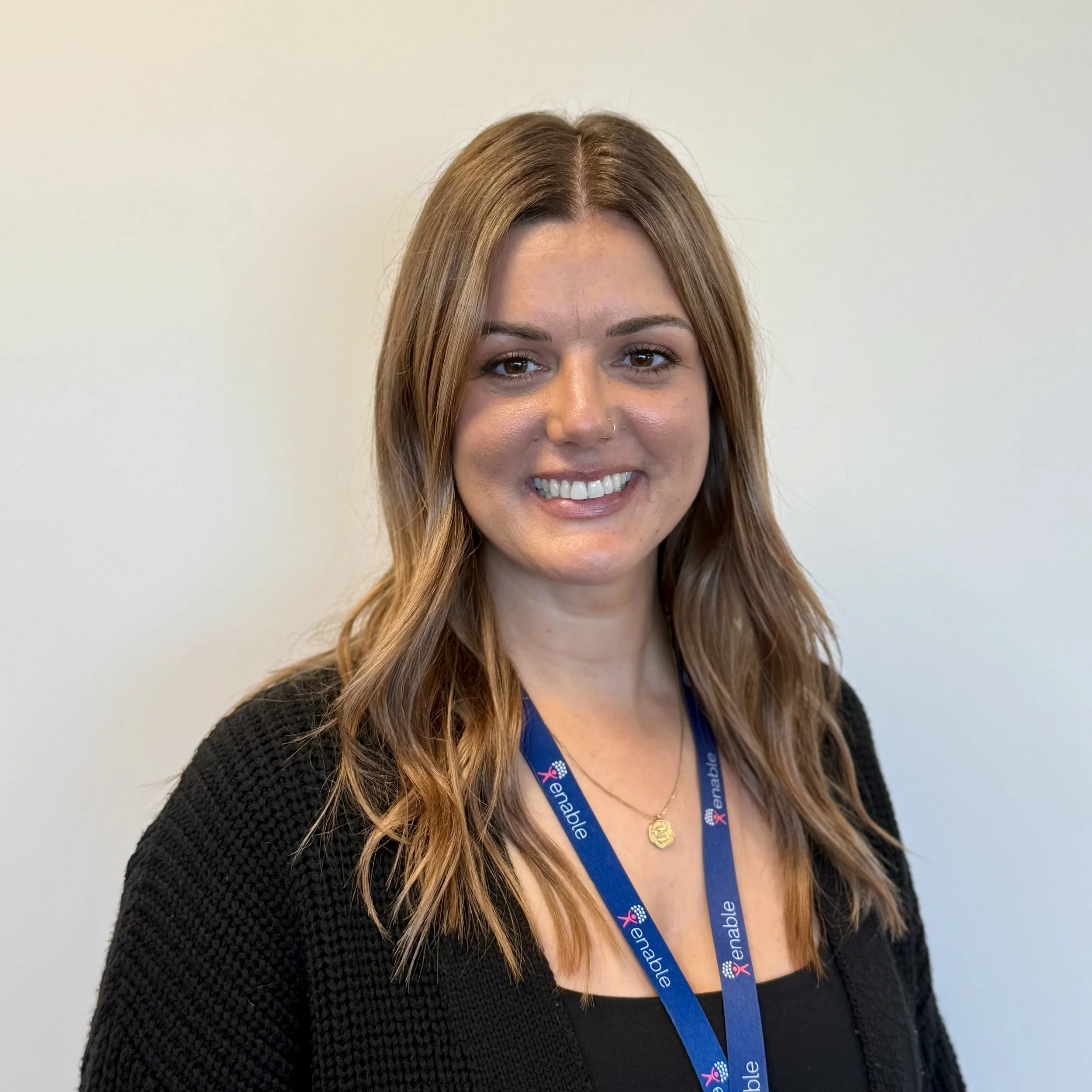 Woman with long brown hair wearing a black shirt and a blue lanyard labeled "Enable," smiling against a plain background.