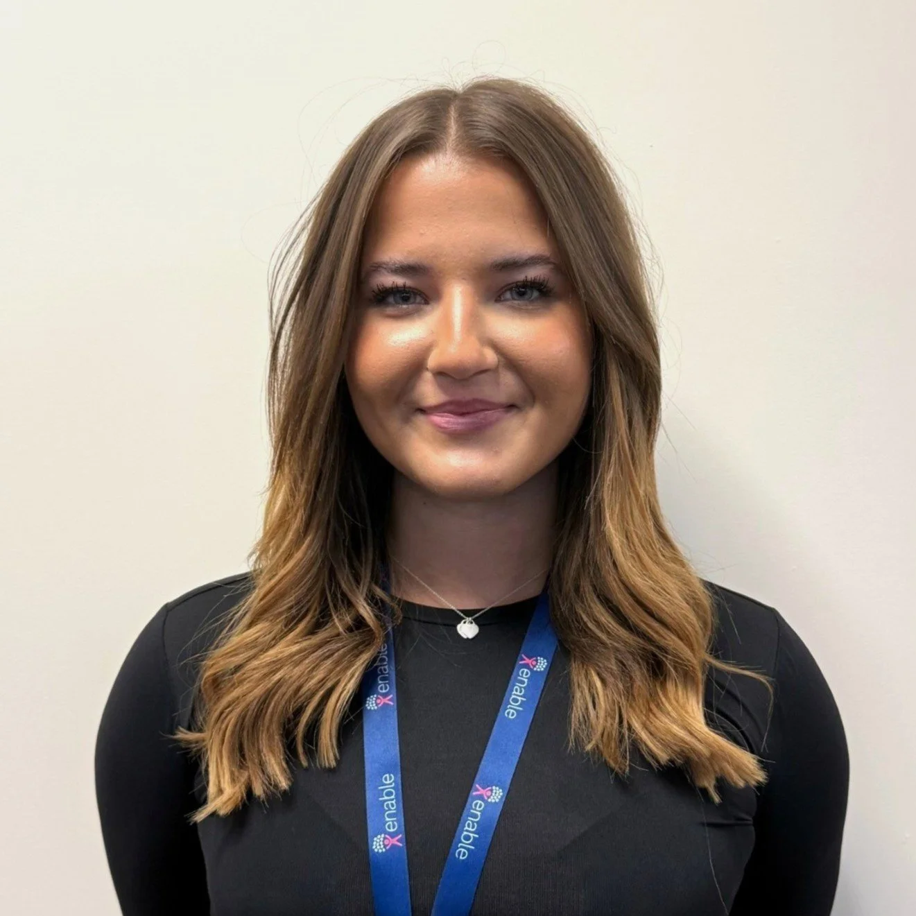 Woman smiling with short light brown hair wearing a black shirt and a lanyard with the text "Enable" against a plain background.