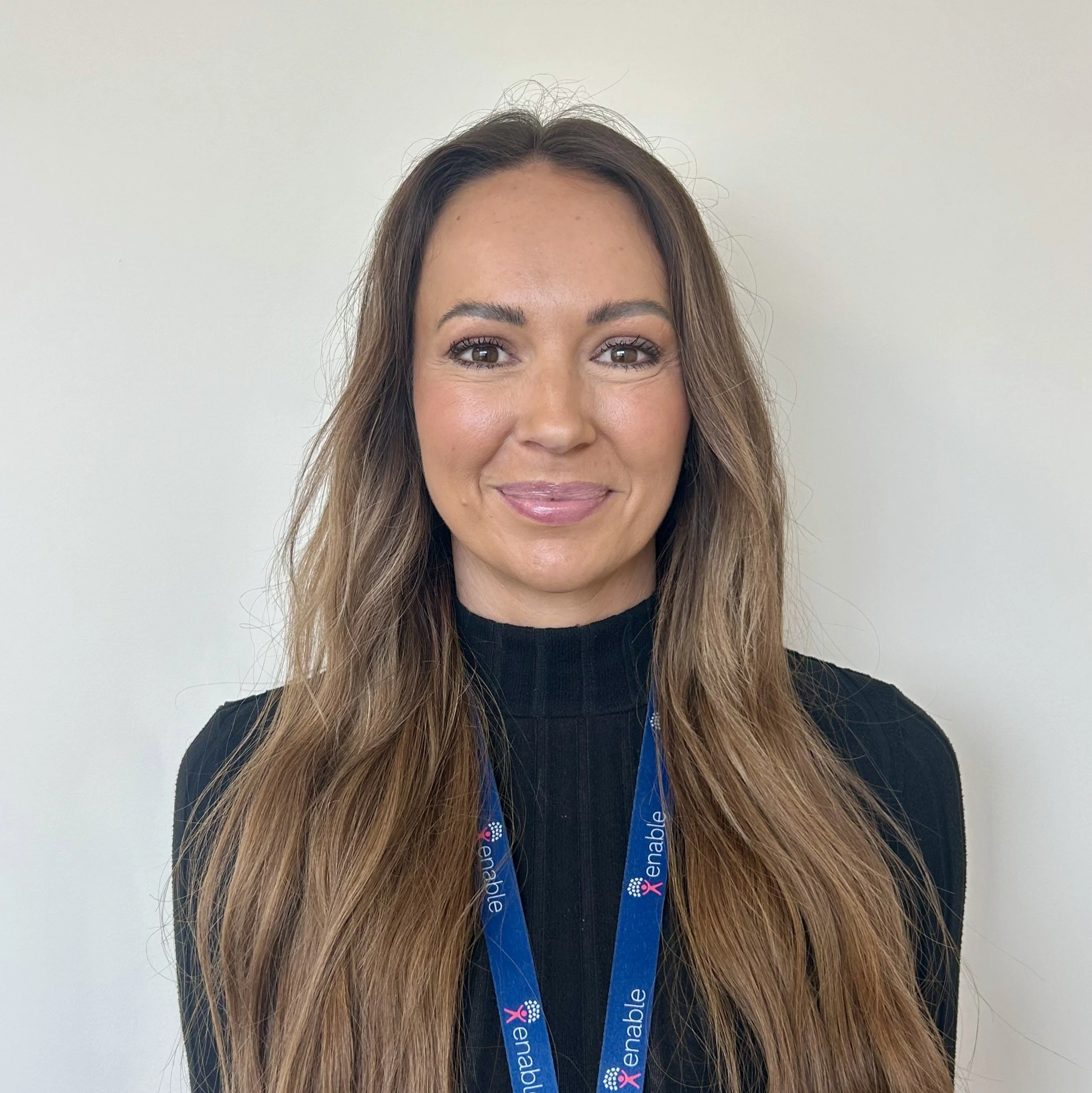 Woman smiling with long brown hair wearing a black top and blue 'Enable' lanyard.