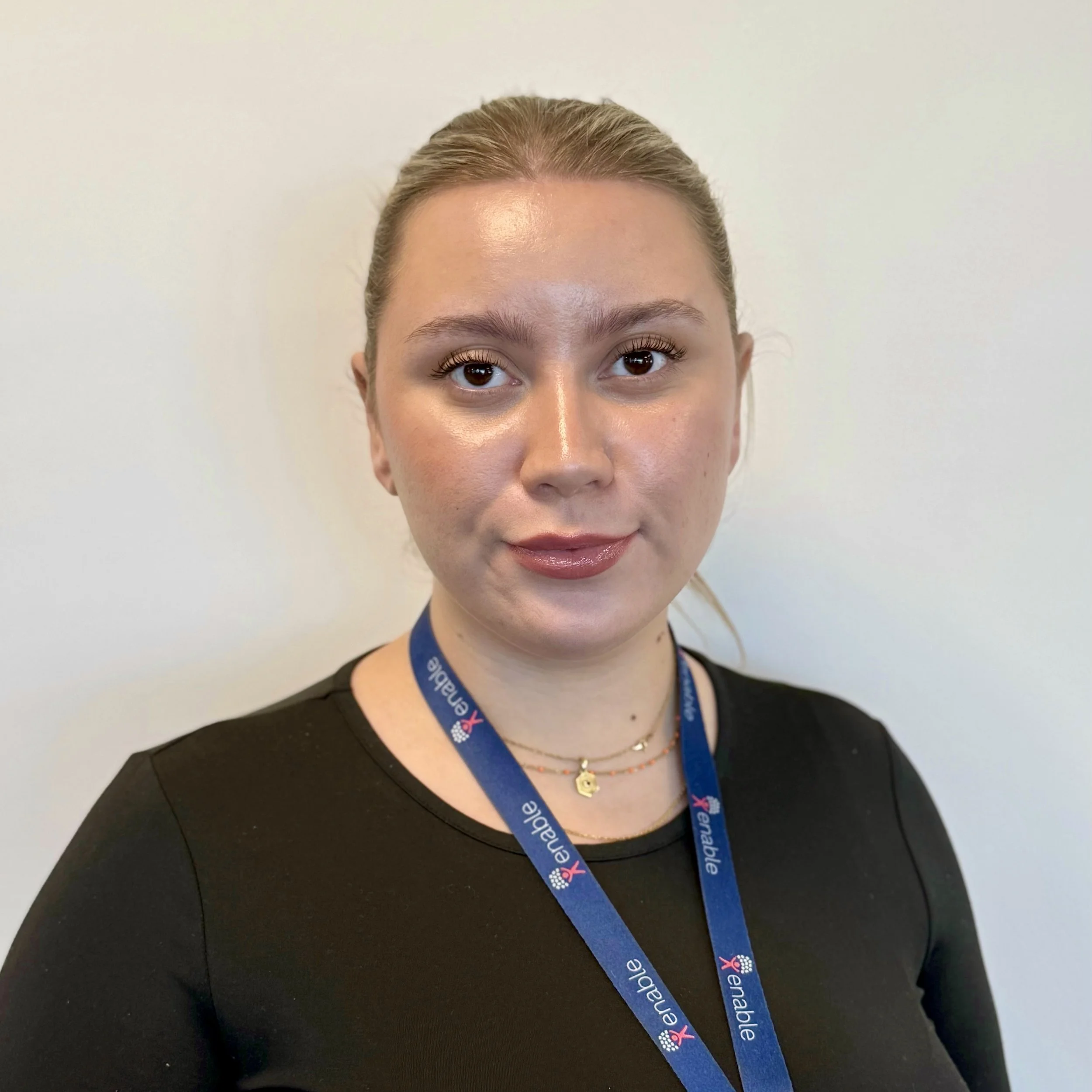 Woman with blonde hair tied up smiling, wearing a black shirt and a blue lanyard, in front of a plain wall.