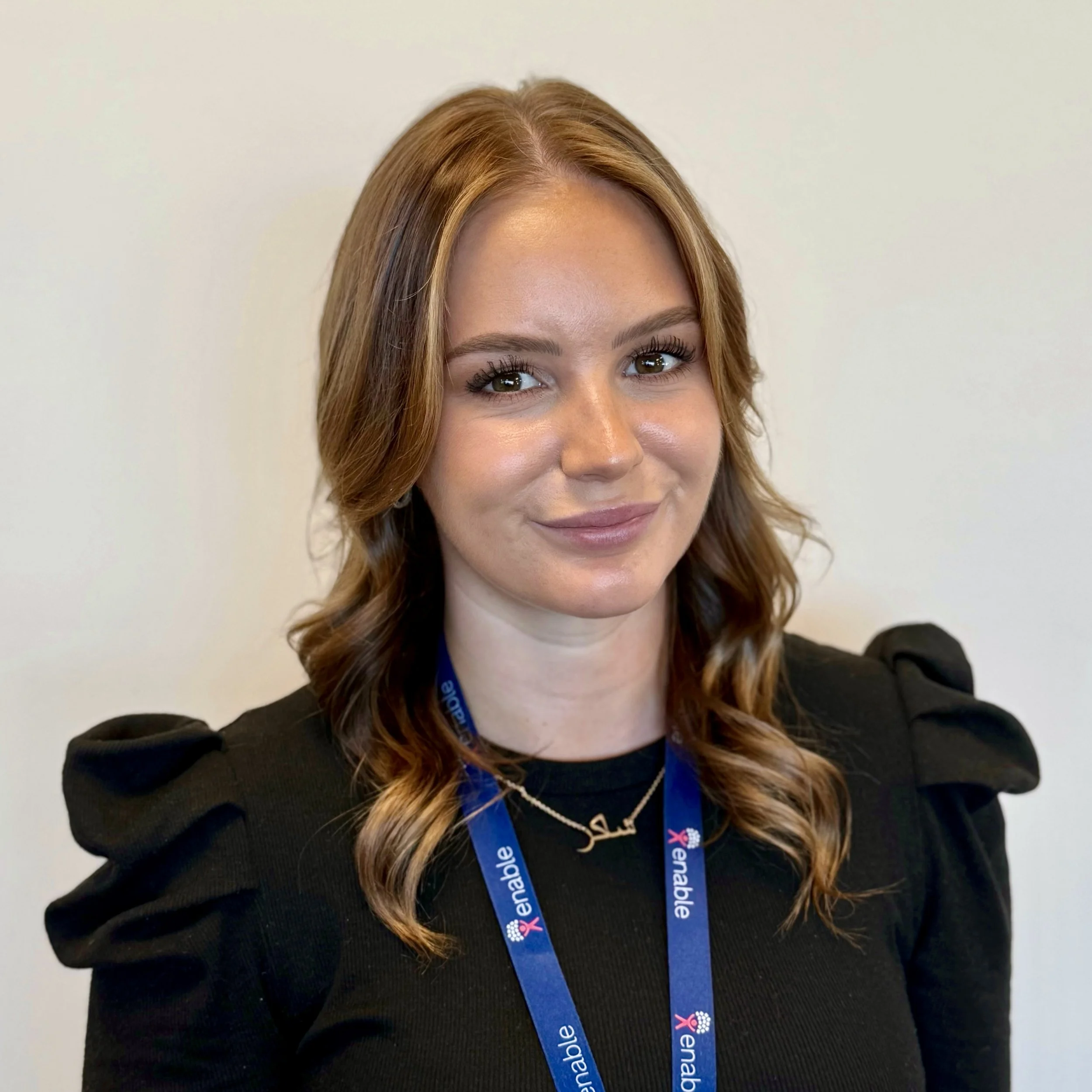 A woman with short brown hair wearing a black top and a blue lanyard, standing against a neutral background.