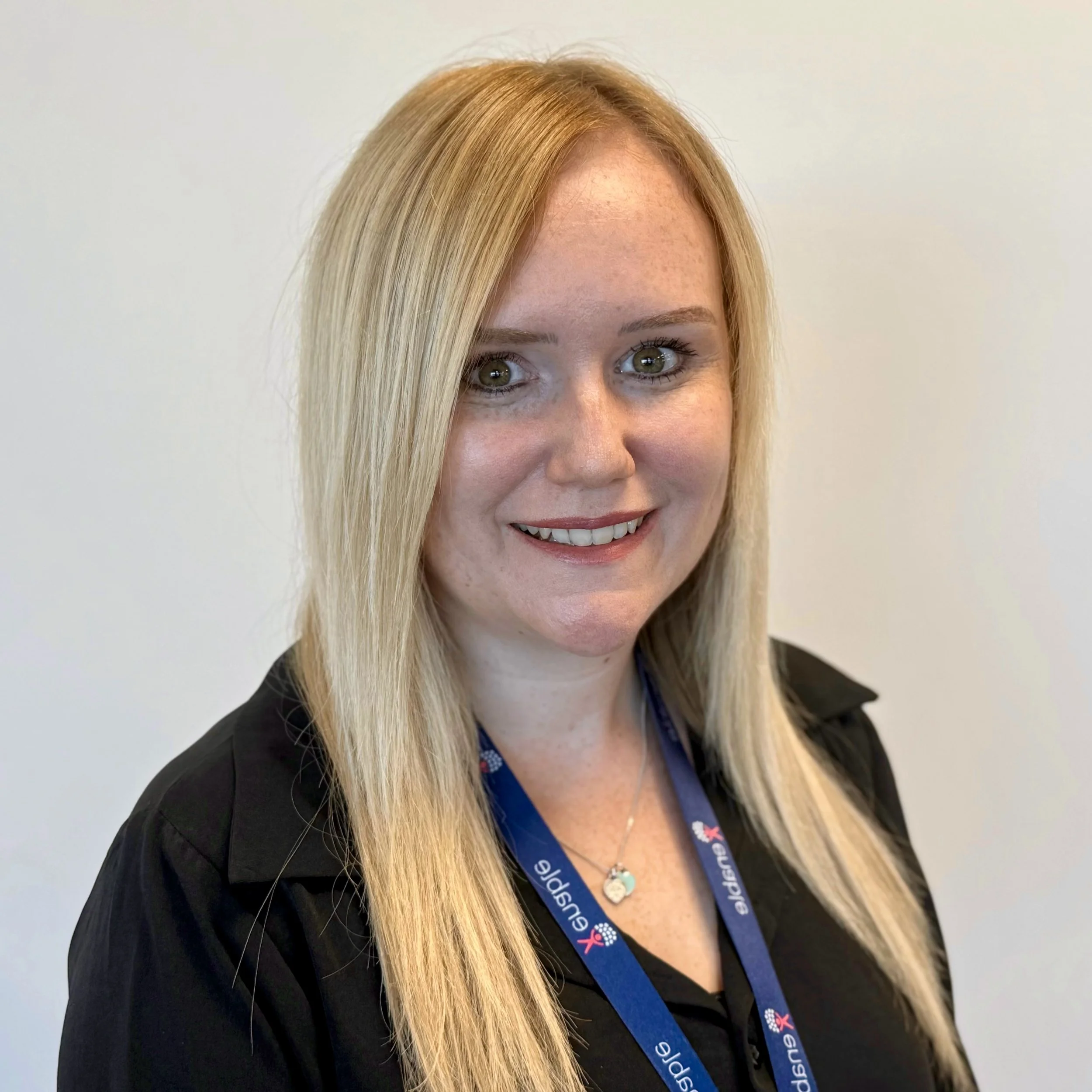 Woman smiling wearing a dark shirt and blue lanyard with the word 'Enable' printed on it, smiling in front of a plain background.