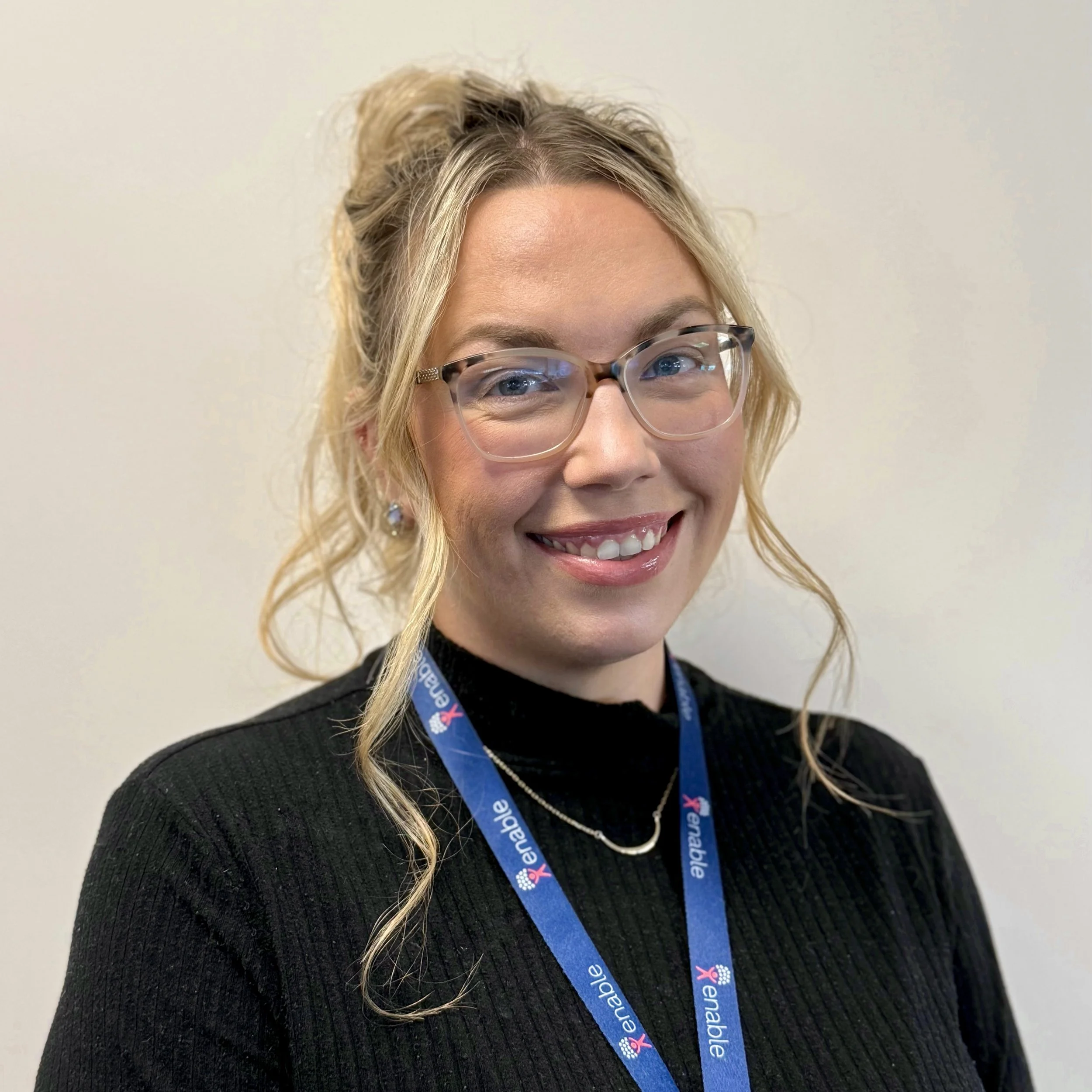 Woman with long blonde hair tied up, wearing glasses and a black top, smiling, with a lanyard that says 'Enable'.