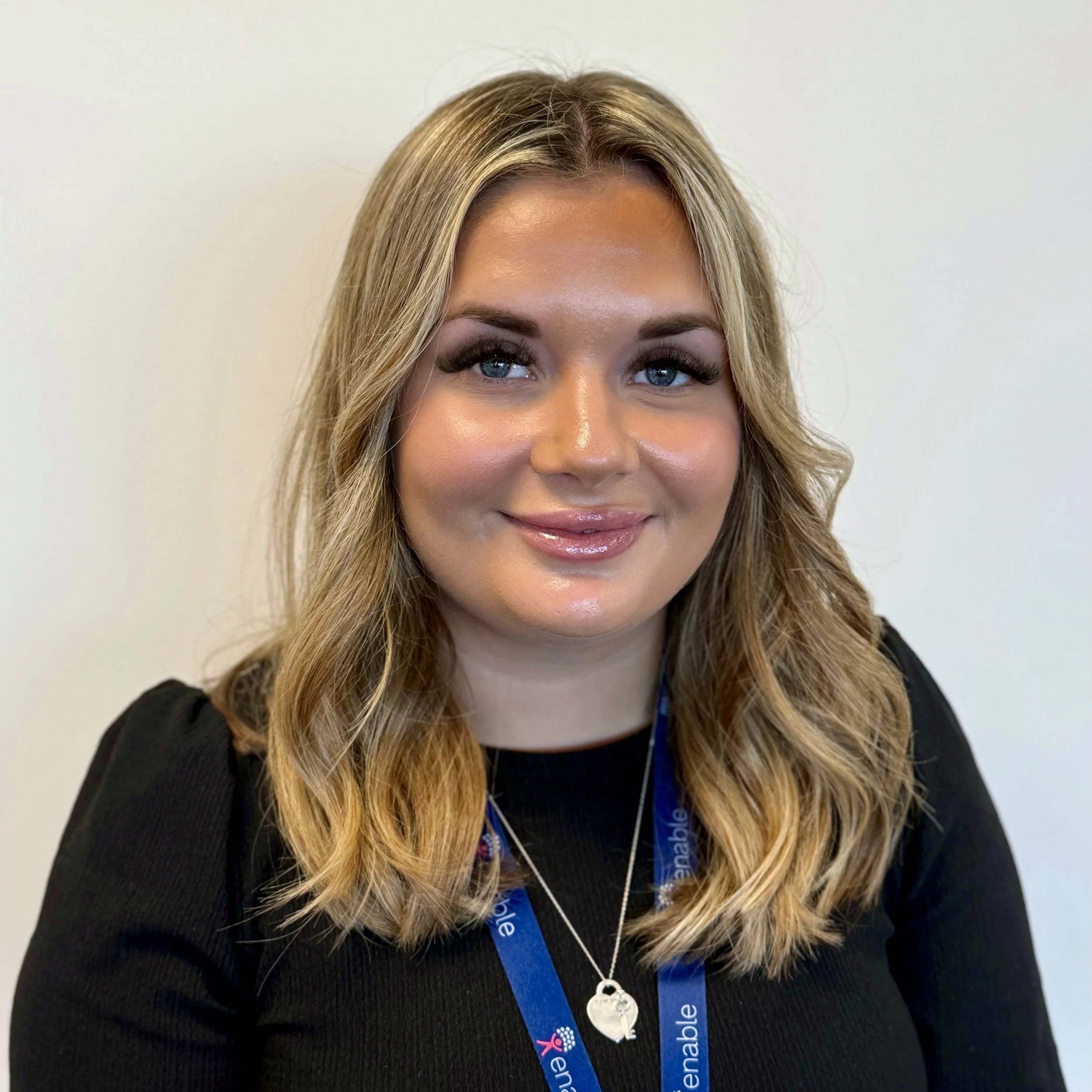 Woman with short light brown hair, wearing a black top and a lanyard, smiling in front of a plain background.