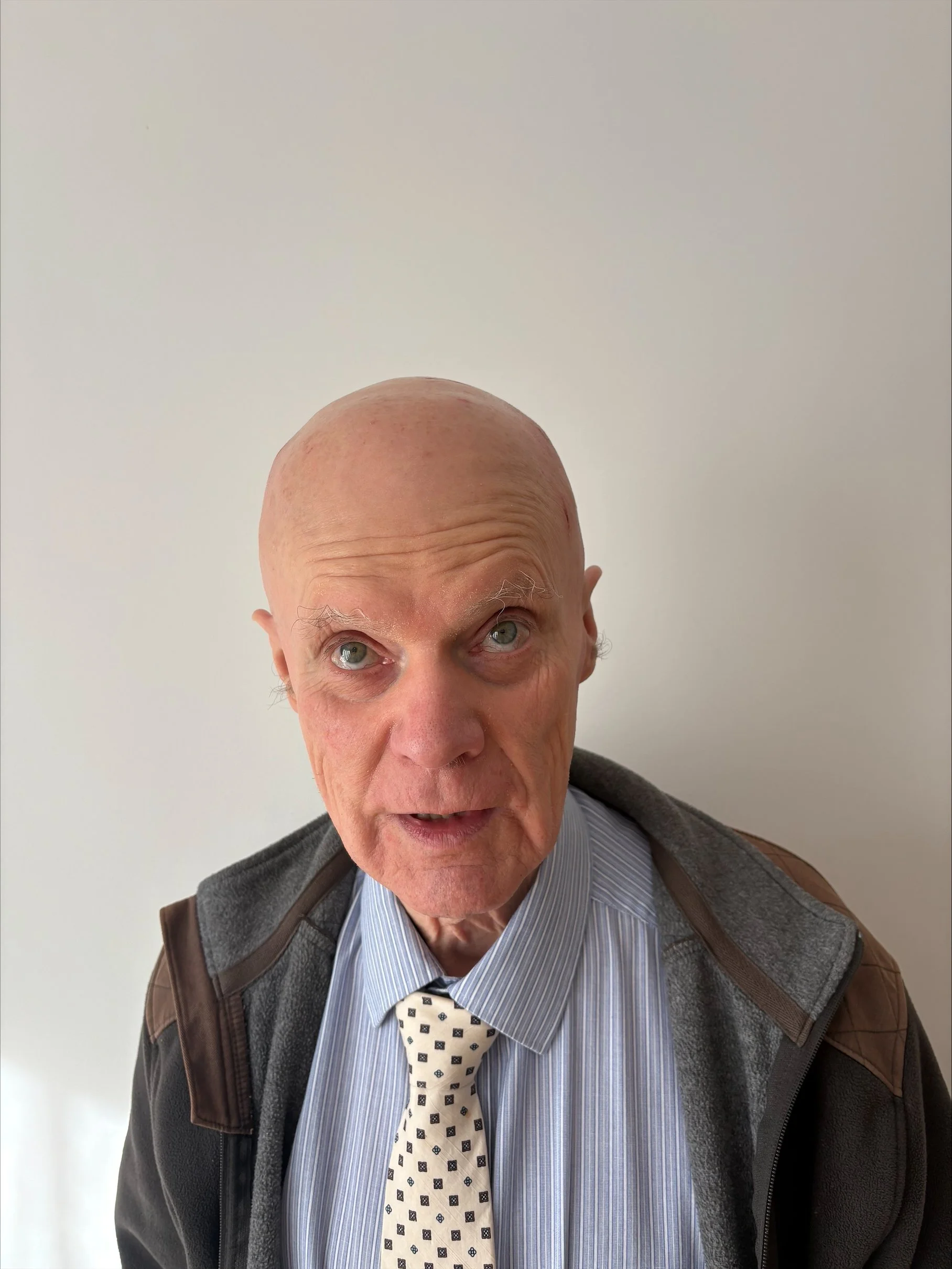 A bald man wearing a smarrt light blue shirt and tie with a jacket, against a plain white wall smiling.