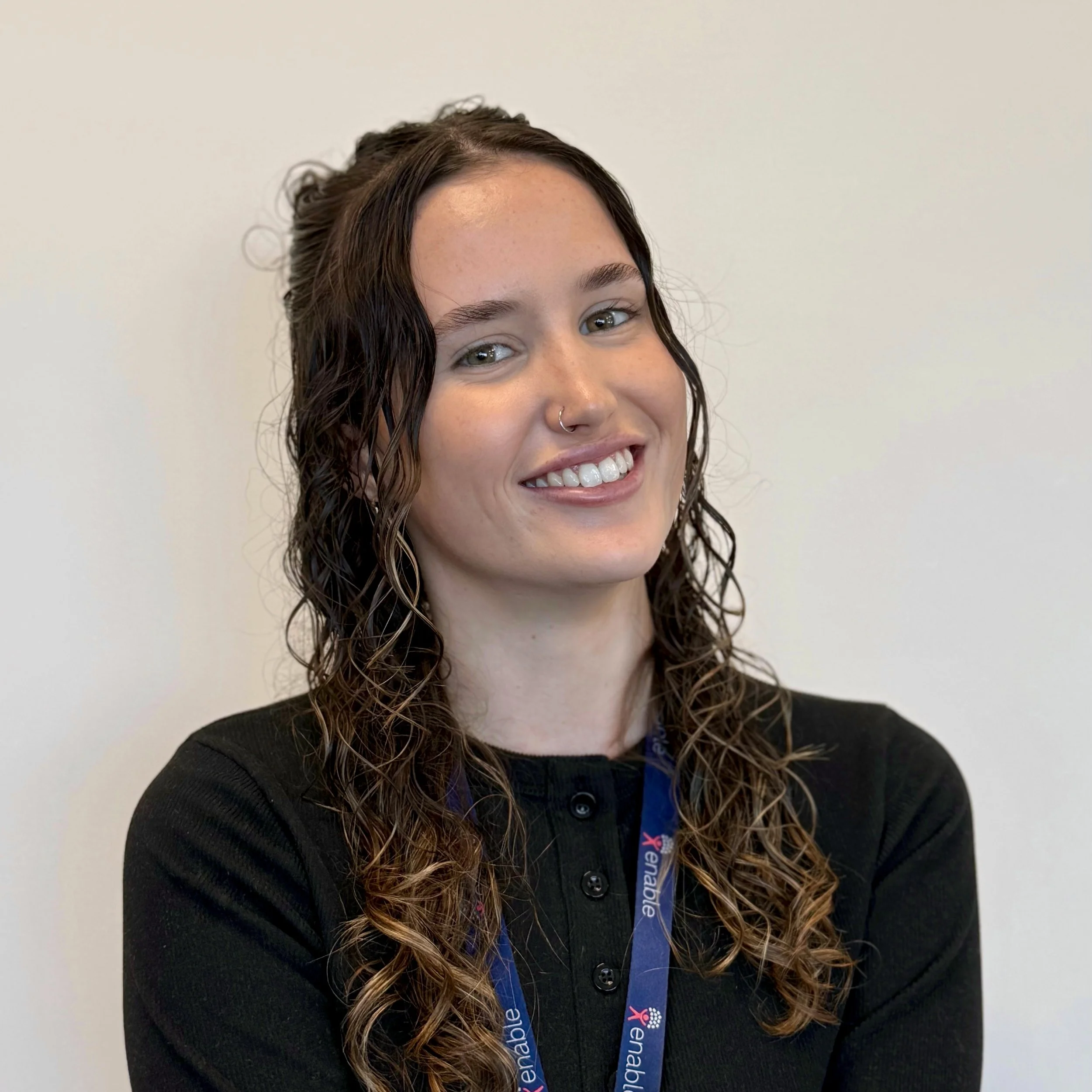 A woman with short brown curly hair, wearing a black top and a lanyard with the text "Enable." They have a necklace with a cross pendant and a nose piercing, standing against a plain background.